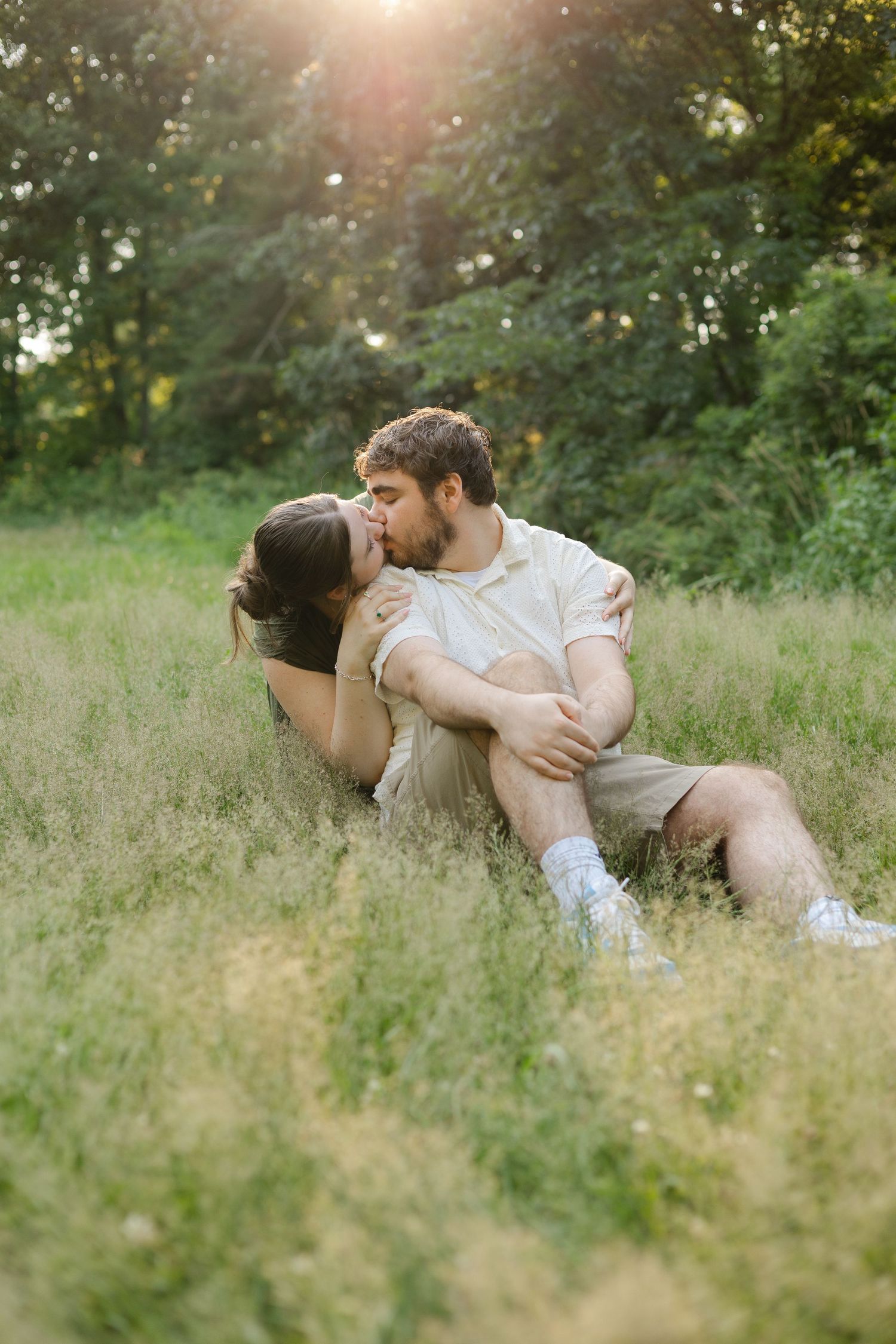 Couple kissing and embracing in a sunlit grassy field surrounded by lush green trees during golden hour.