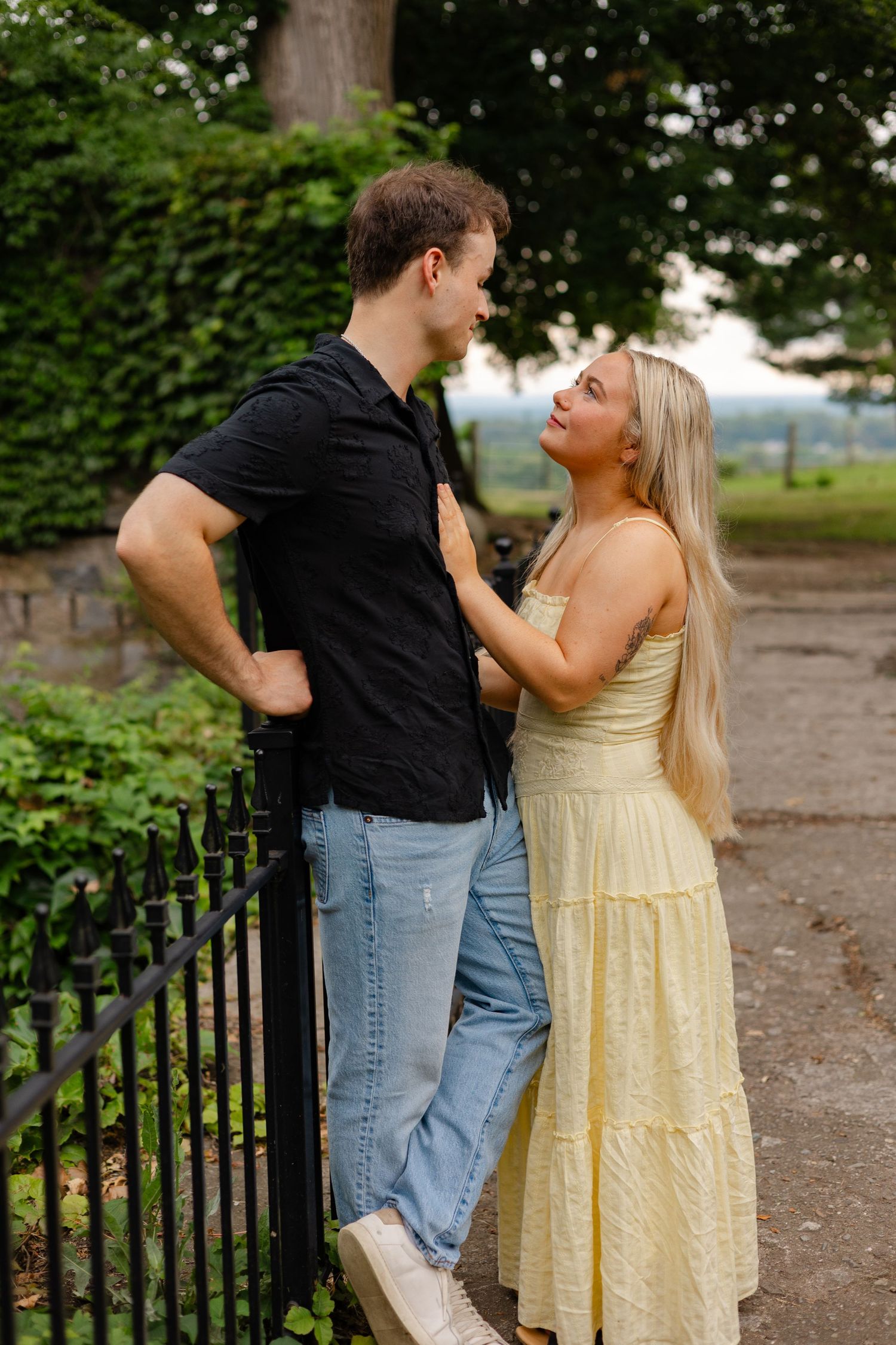 A couple shares a romantic moment by a black iron fence, surrounded by lush greenery outdoors.