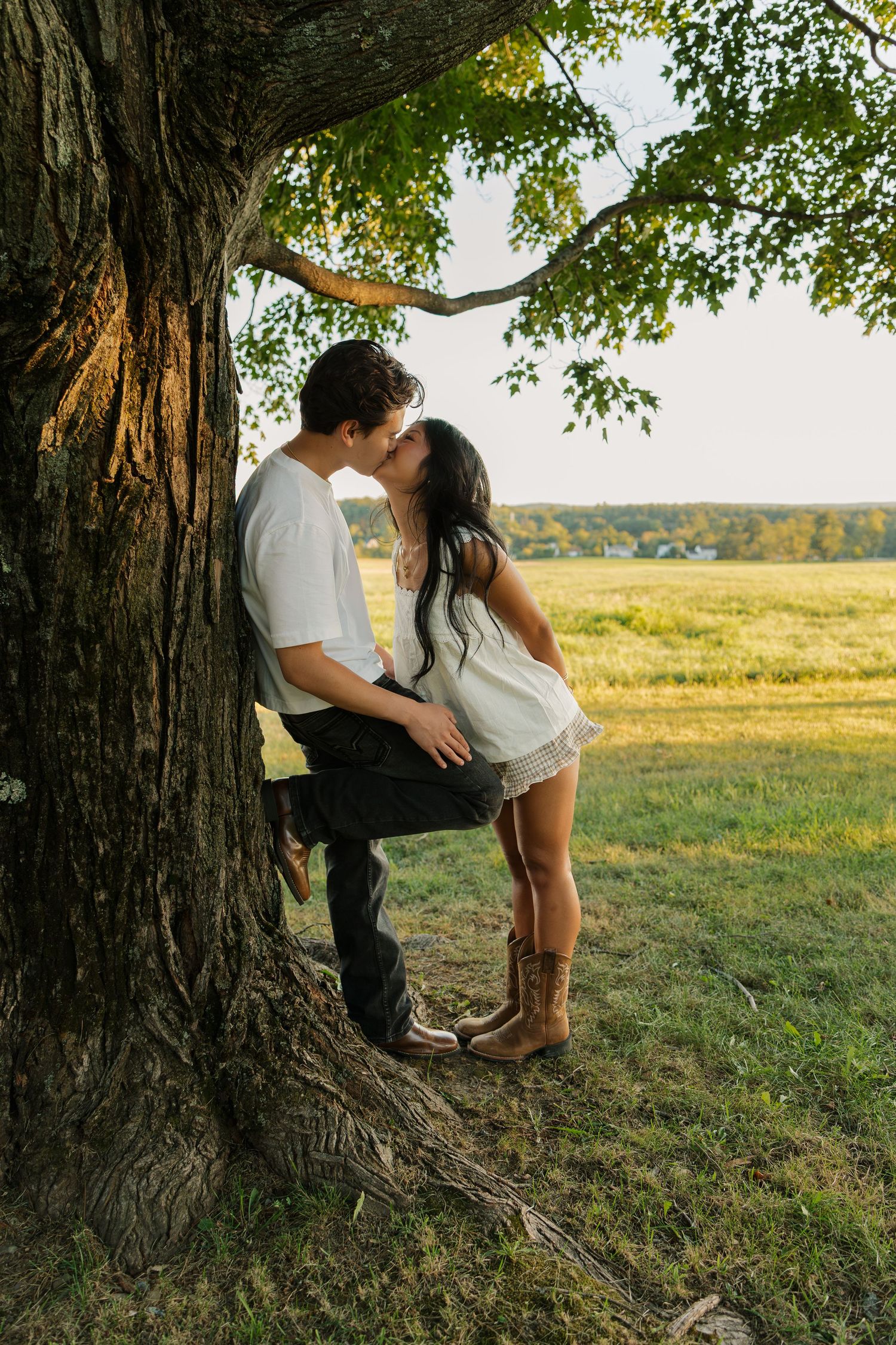 Couple kissing under a large tree in a sunny field, woman in white dress and cowboy boots, man in white shirt.