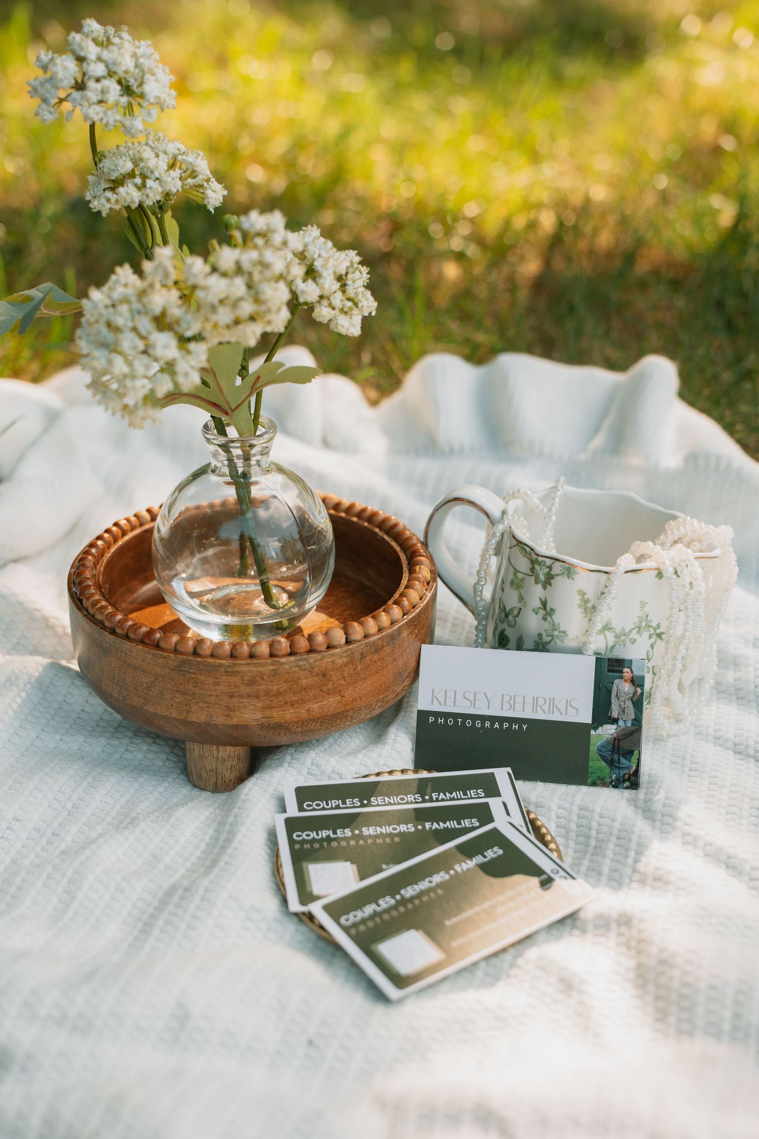 Photography business cards displayed on a white blanket with wildflowers in a glass vase and floral mug outdoors.