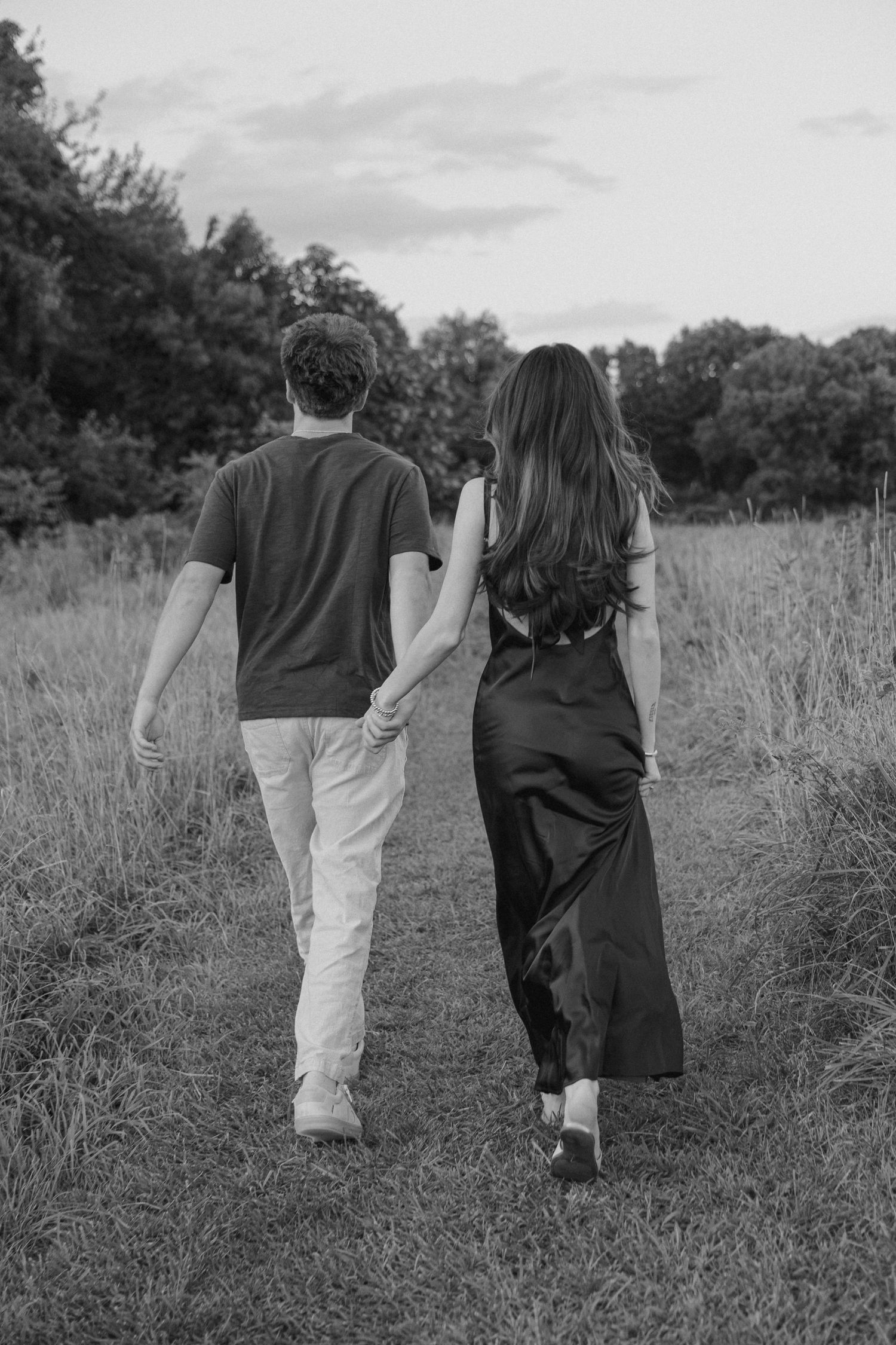 Black and white photo of a couple holding hands while walking along a rural path surrounded by tall grass and trees.
