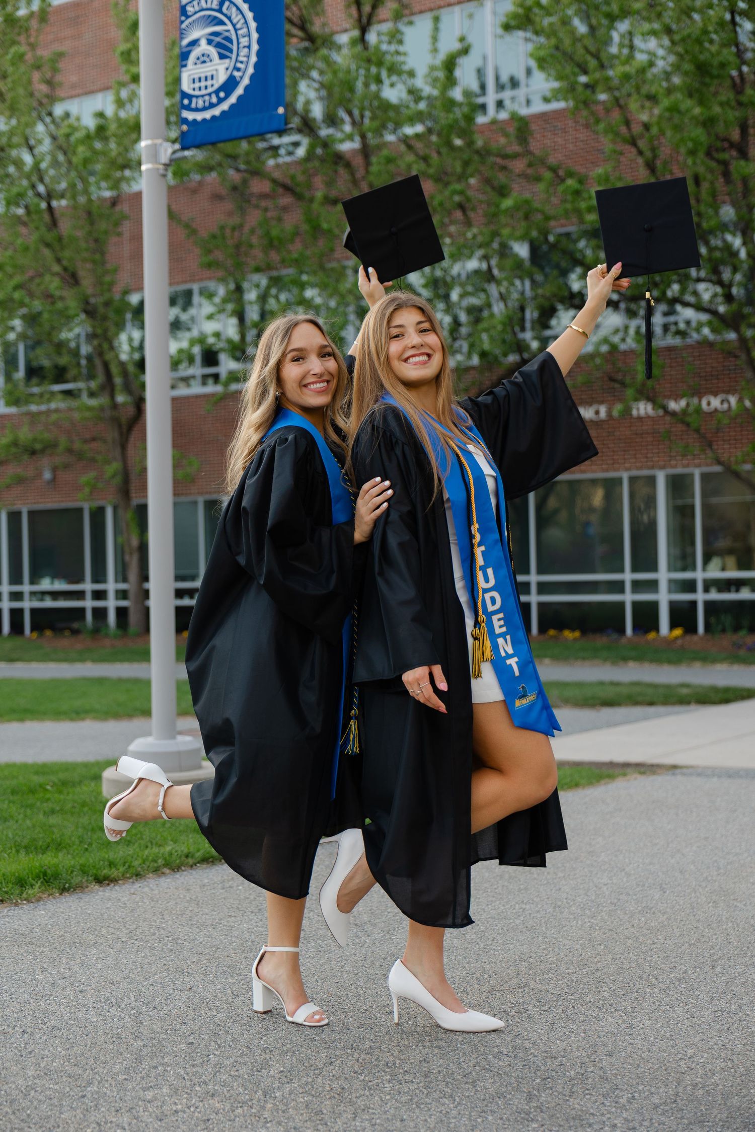 Two smiling women in black graduation gowns and caps celebrate outdoors on a college campus.