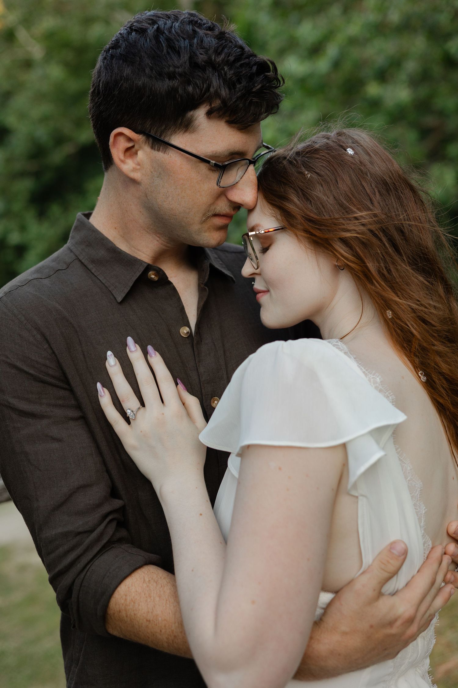 A couple in glasses shares a tender forehead-to-forehead moment, bride in white dress and groom in dark shirt outdoors.
