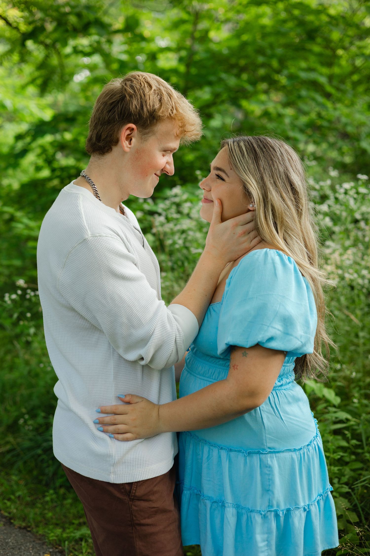 A young couple in a lush green garden, sharing a tender moment, the woman in a blue dress touching the man's face.