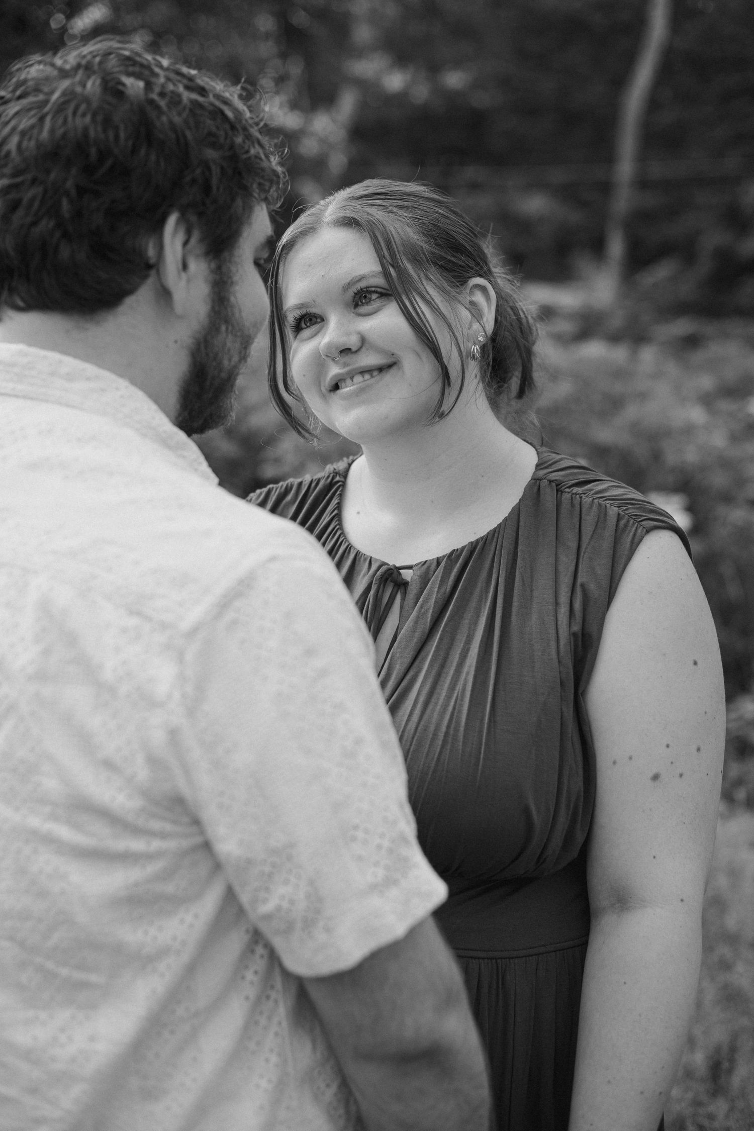 Black and white photo of a smiling woman in a dress gazing at a bearded man outdoors.