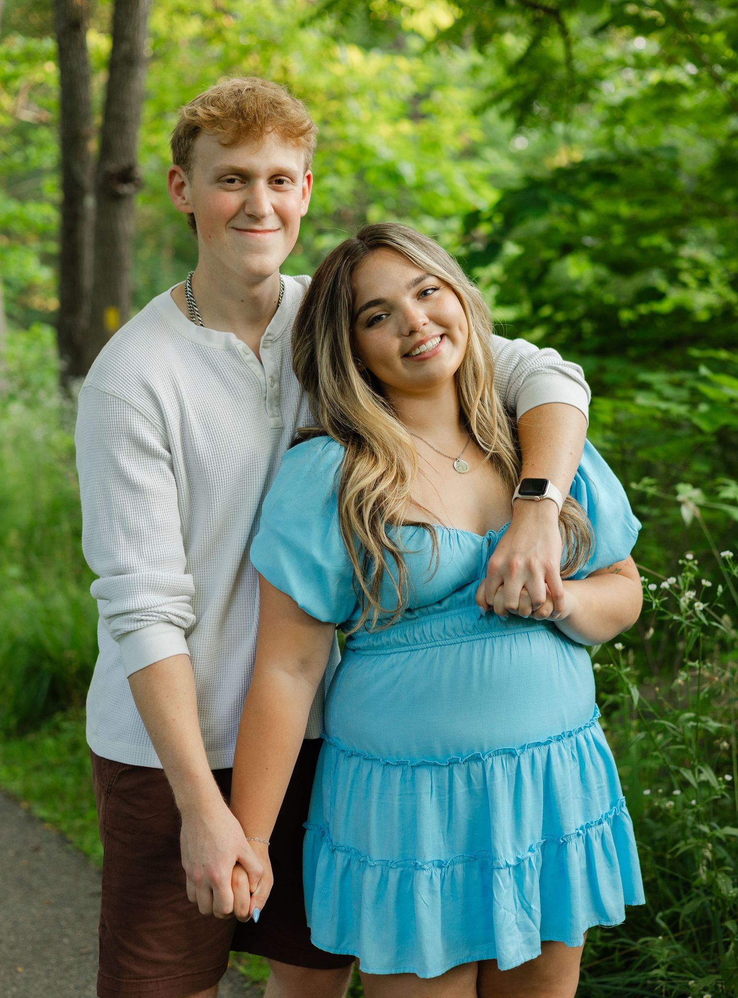A smiling young couple poses together outdoors in a lush green park, wearing casual summer outfits.