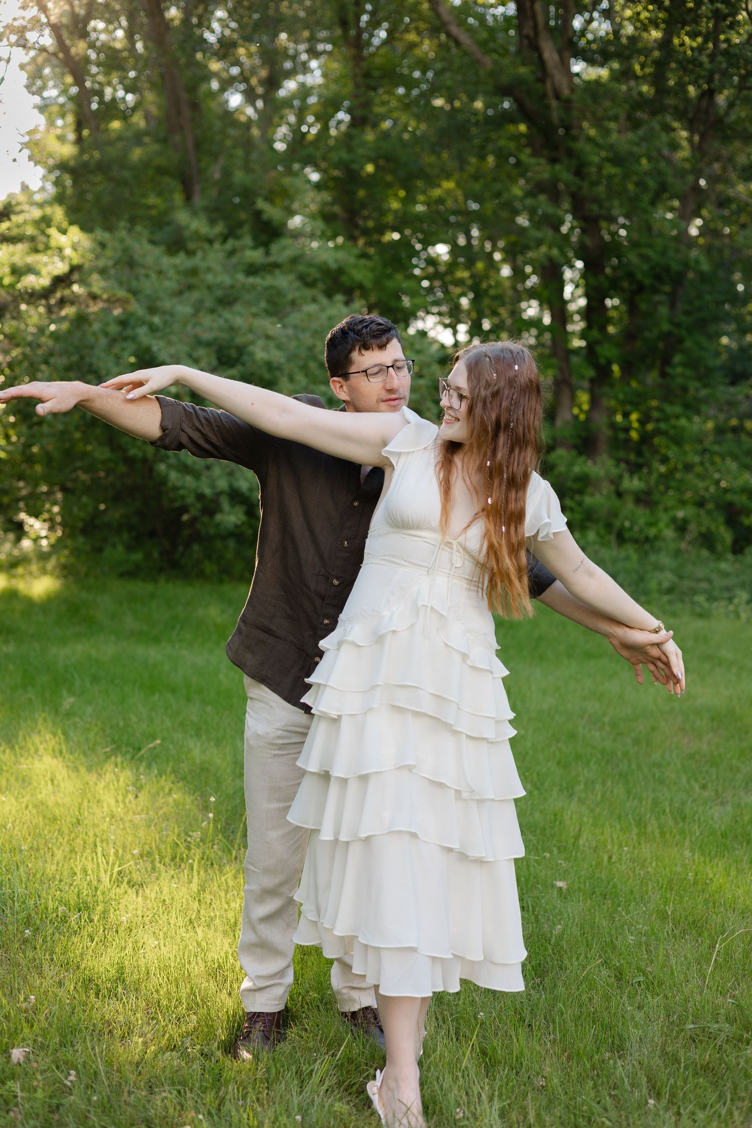 Couple playfully spreading arms like airplane wings in a lush green park, woman in white ruffled dress.