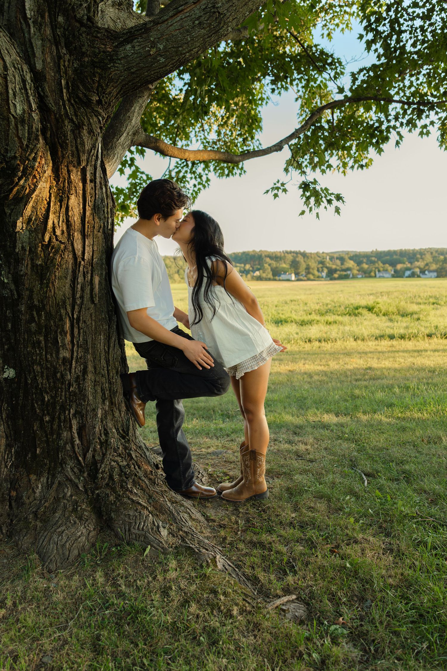 A couple shares a romantic kiss leaning against a large tree in a sunlit green field, woman wearing cowboy boots.