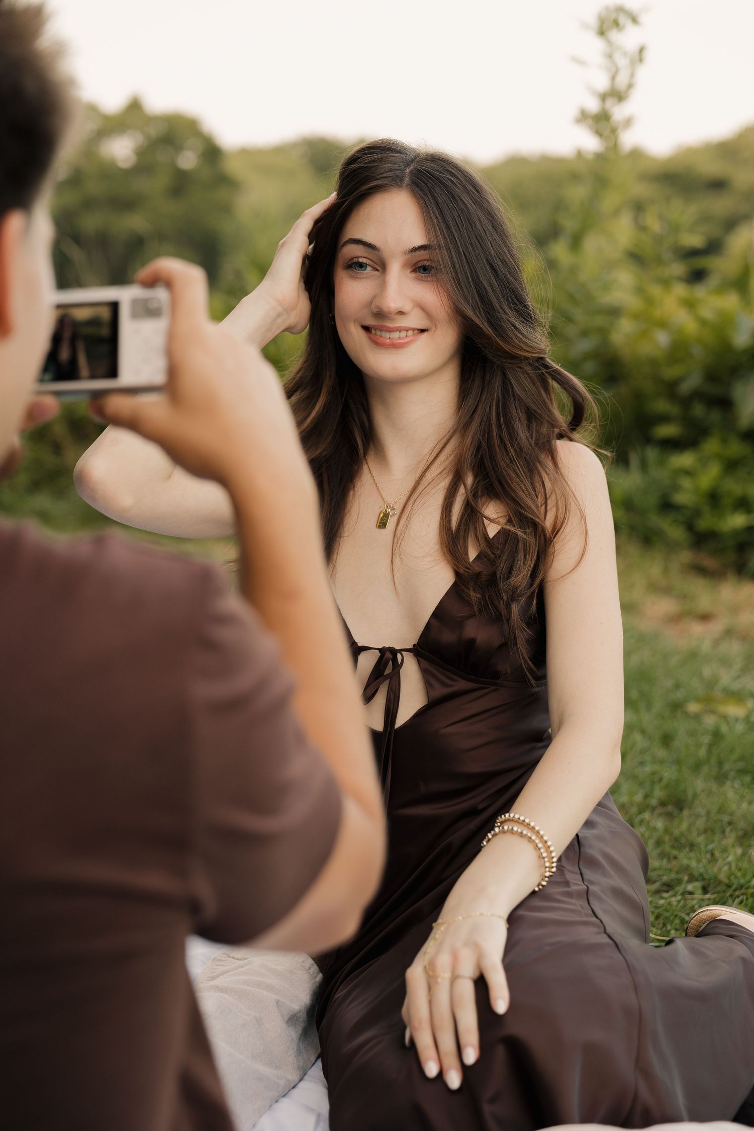 Young woman in brown dress smiling while being photographed outdoors in a lush green park setting.
