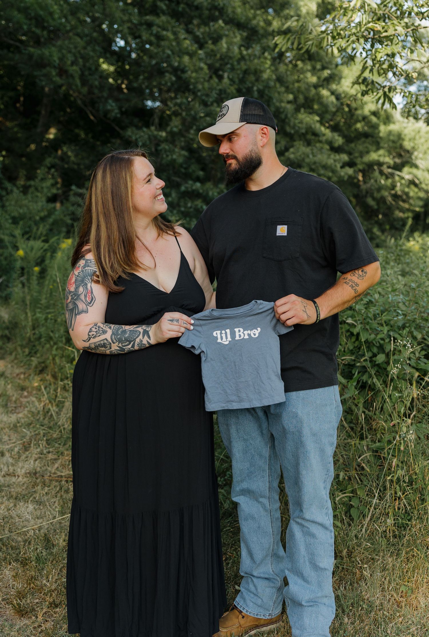 Couple holding a lil bro baby shirt outdoors, announcing pregnancy with a boy, surrounded by lush green trees.