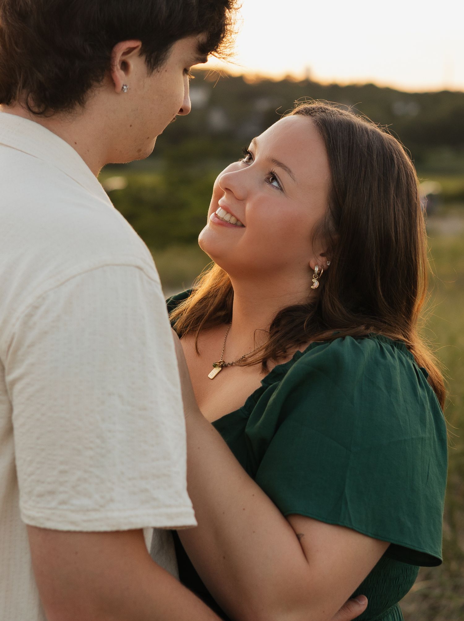 A smiling young woman in a green dress gazes up at a man during a romantic golden hour outdoor photo session.