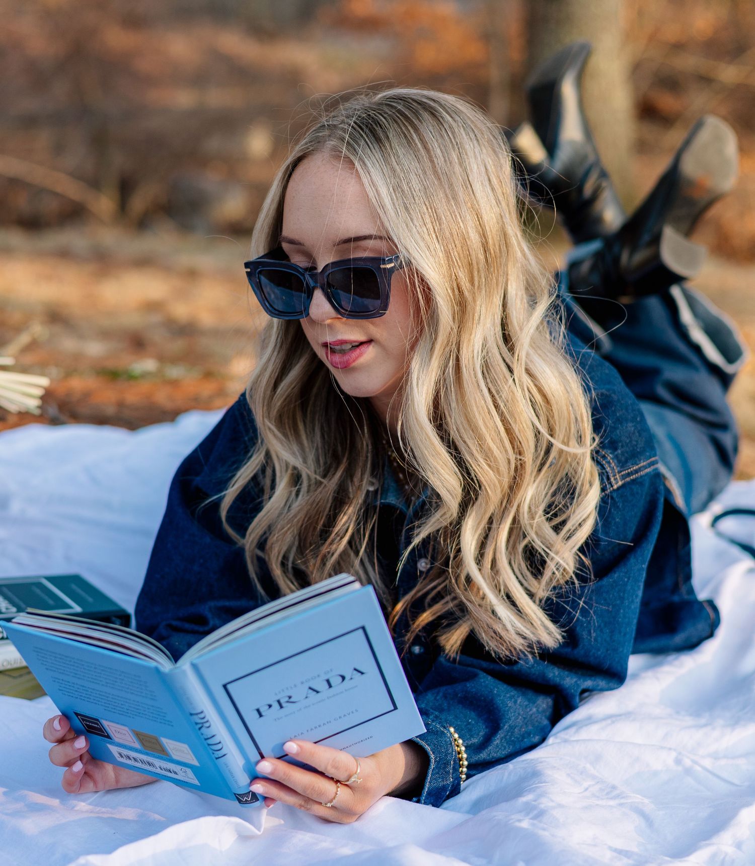 Blonde woman in denim jacket and Prada sunglasses reading a Prada book while lying on a white blanket outdoors.