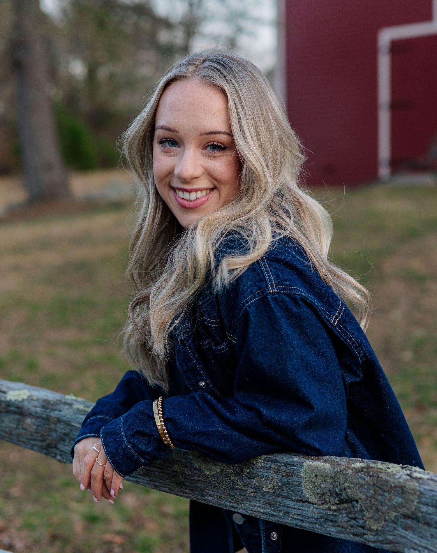 Young blonde woman in denim jacket smiling outdoors by rustic wooden fence with red barn in background.
