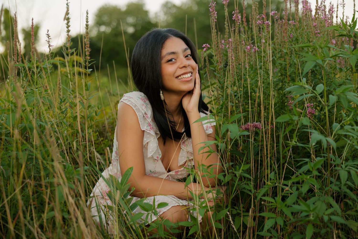 A young woman smiles warmly while crouching among tall wildflowers and green plants in a lush outdoor field.
