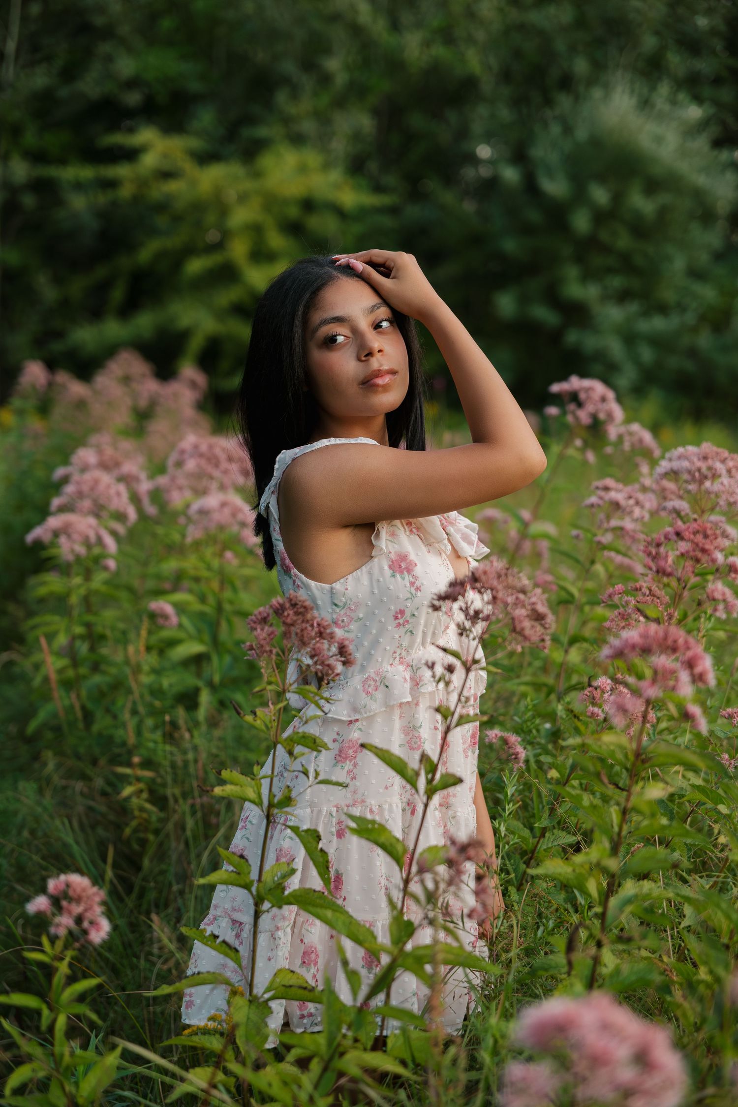 Young woman in floral dress posing among pink wildflowers in a lush green garden setting.