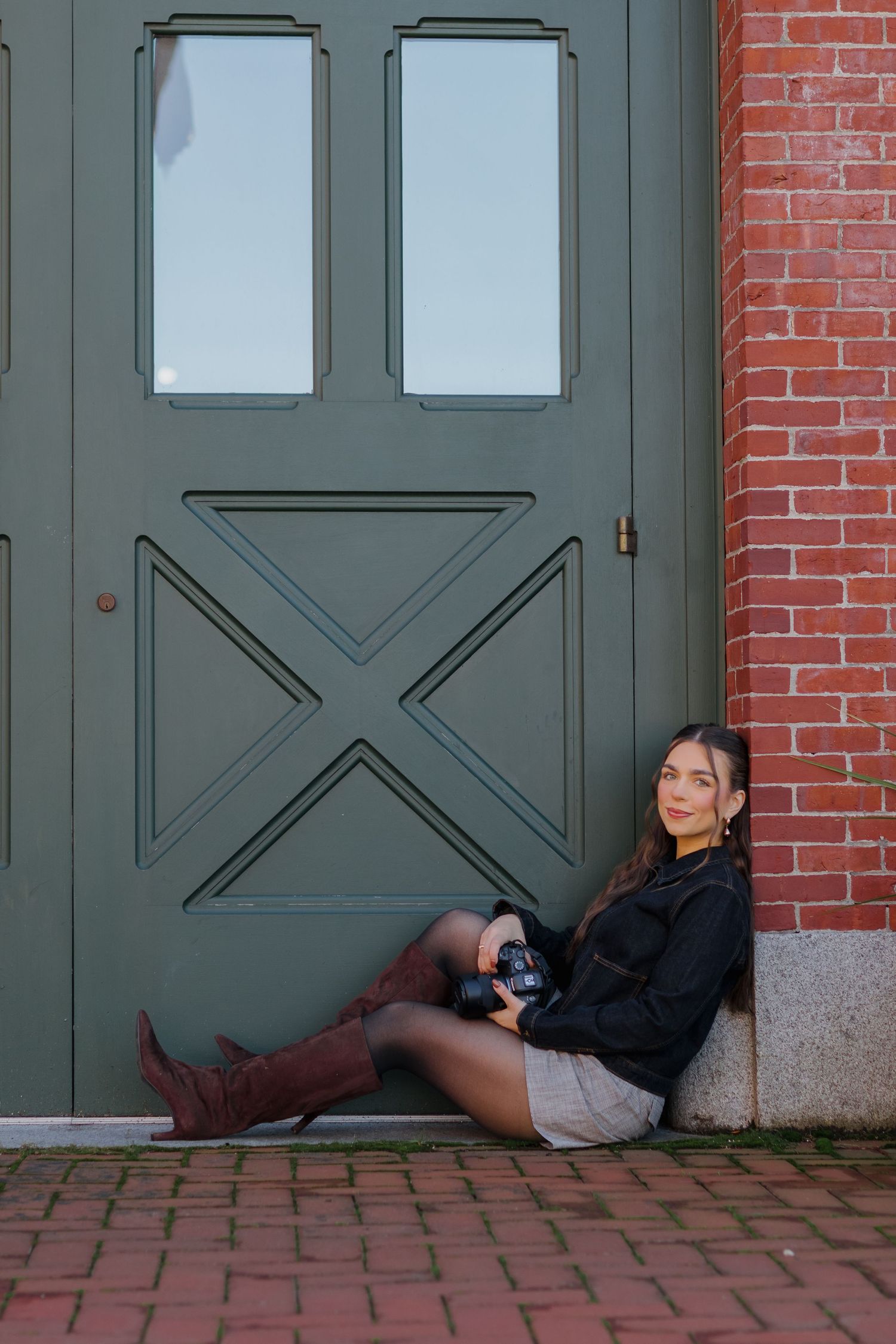 Woman in a black jacket and brown knee-high boots sits against a green paneled door and red brick wall, holding a camera.