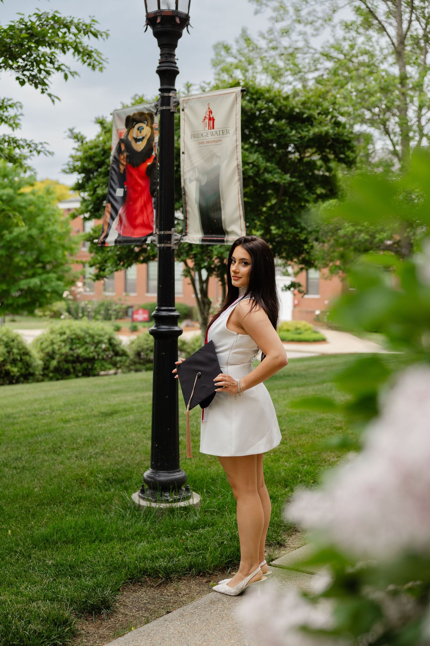 Young woman in white dress holding graduation cap beside a lamp post on a college campus.