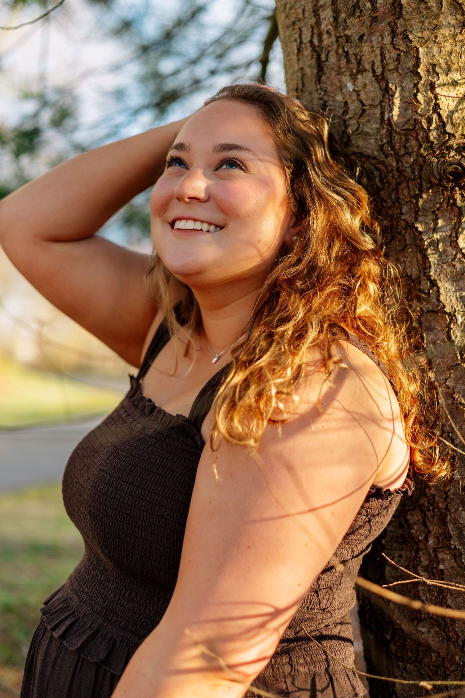 Curly-haired woman in brown sundress smiling while leaning against tree in golden sunset light.