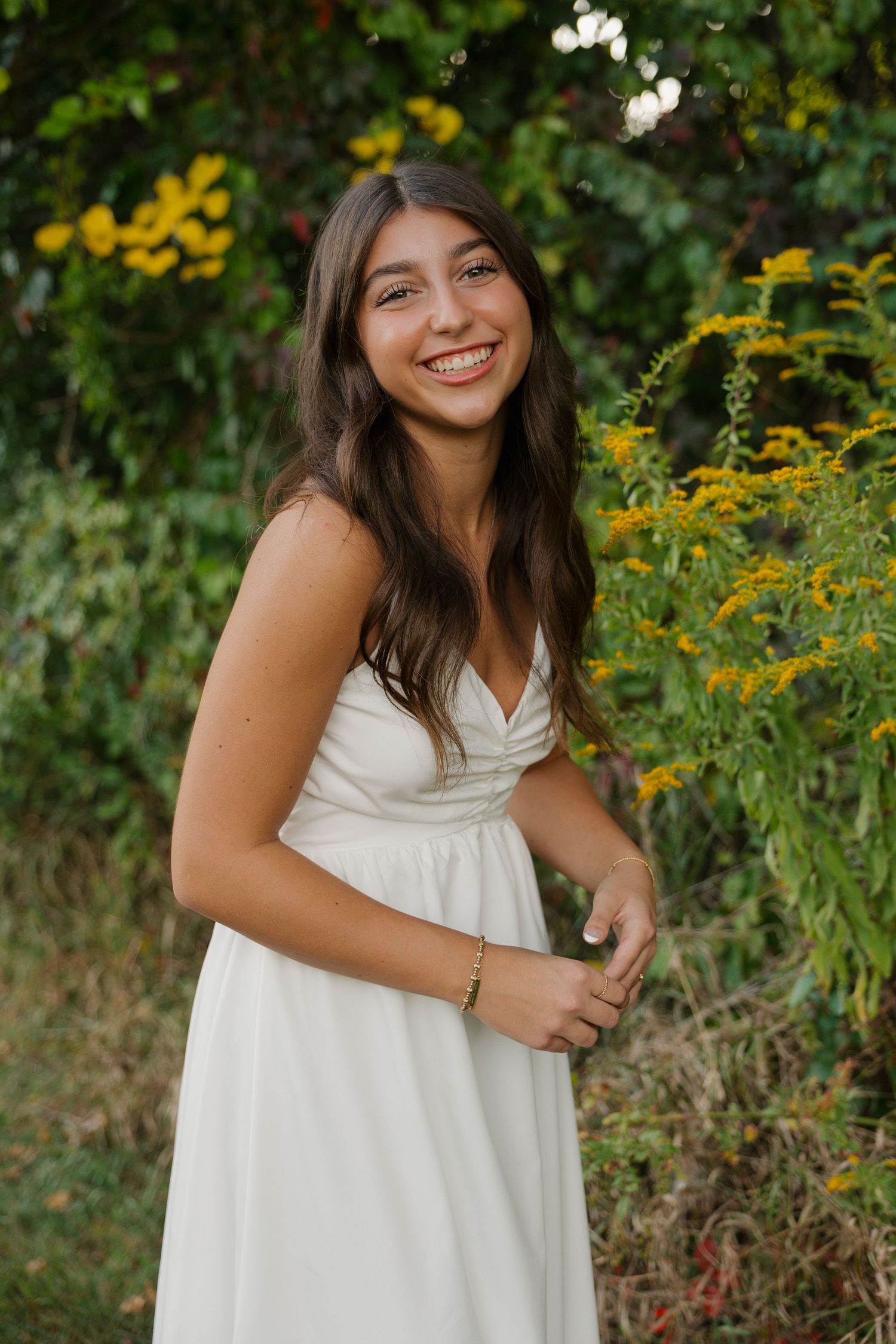 Young woman in white sundress smiling outdoors among yellow wildflowers and green foliage.