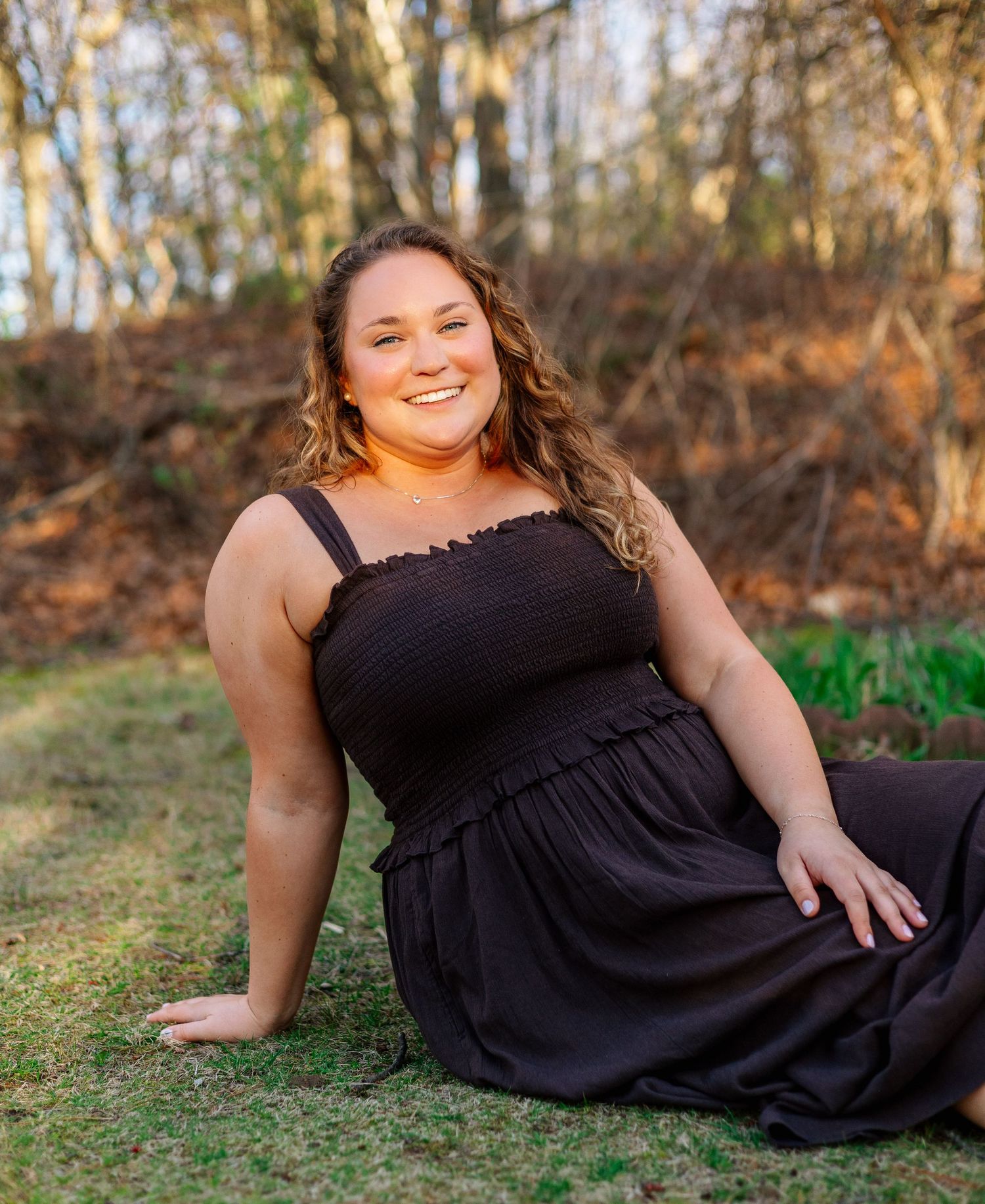 Young woman in black smocked sundress sitting on grass, smiling outdoors in golden hour light among trees.