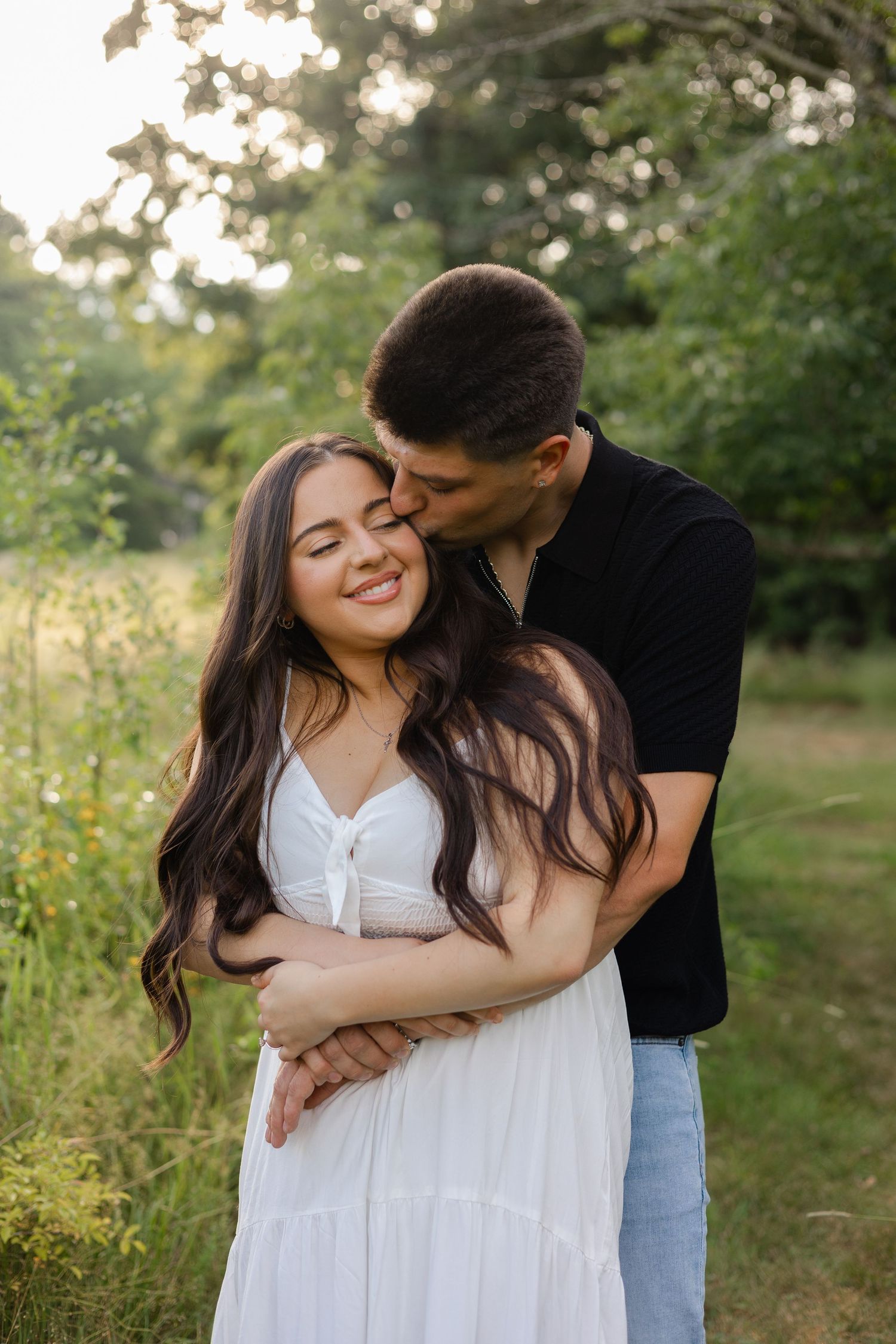 A couple embraces outdoors, woman in white dress smiling as man in black shirt kisses her cheek in a lush green field.