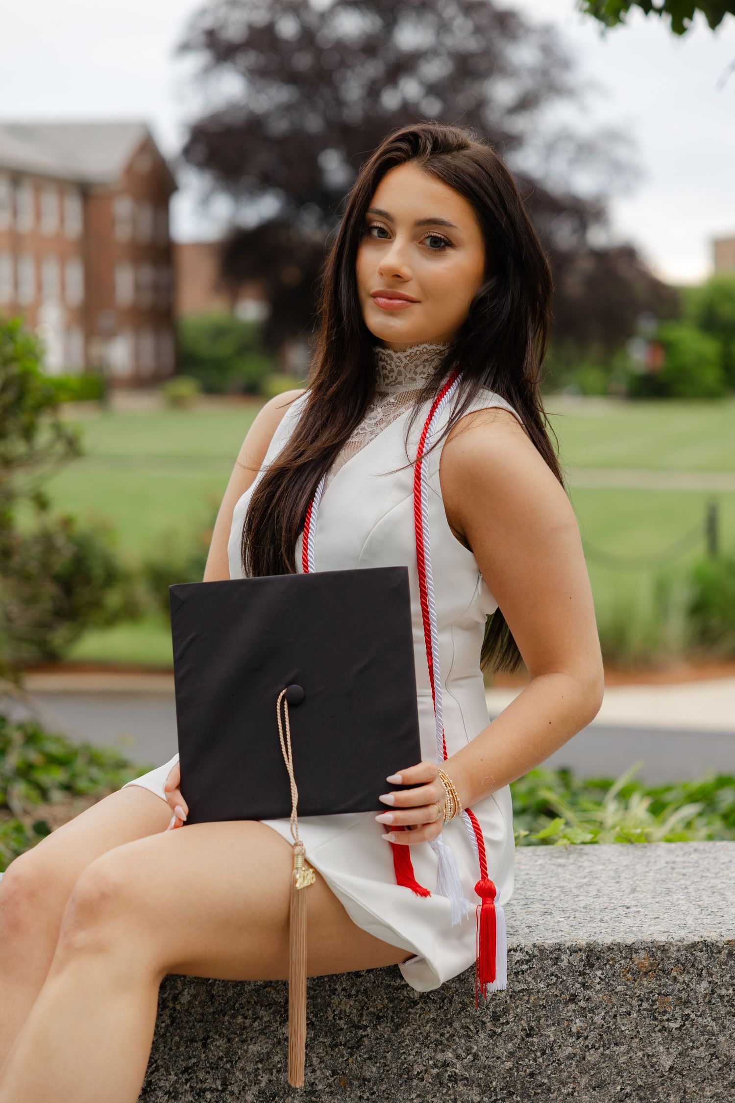 Young woman in white dress holding graduation cap with red tassel, posing outdoors on campus grounds.