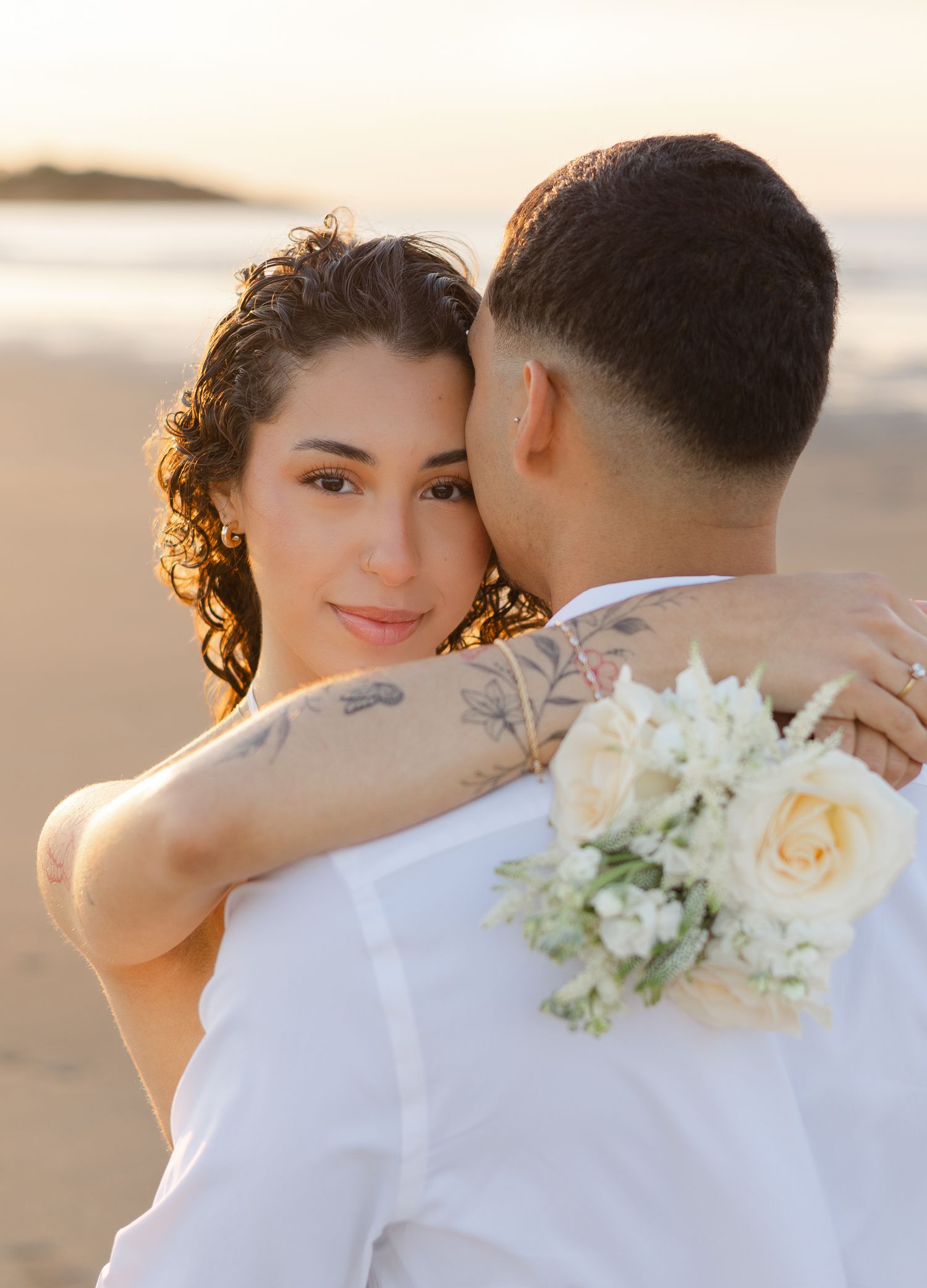 Woman with curly hair and arm tattoo embracing man while holding white rose bouquet on beach at sunset.