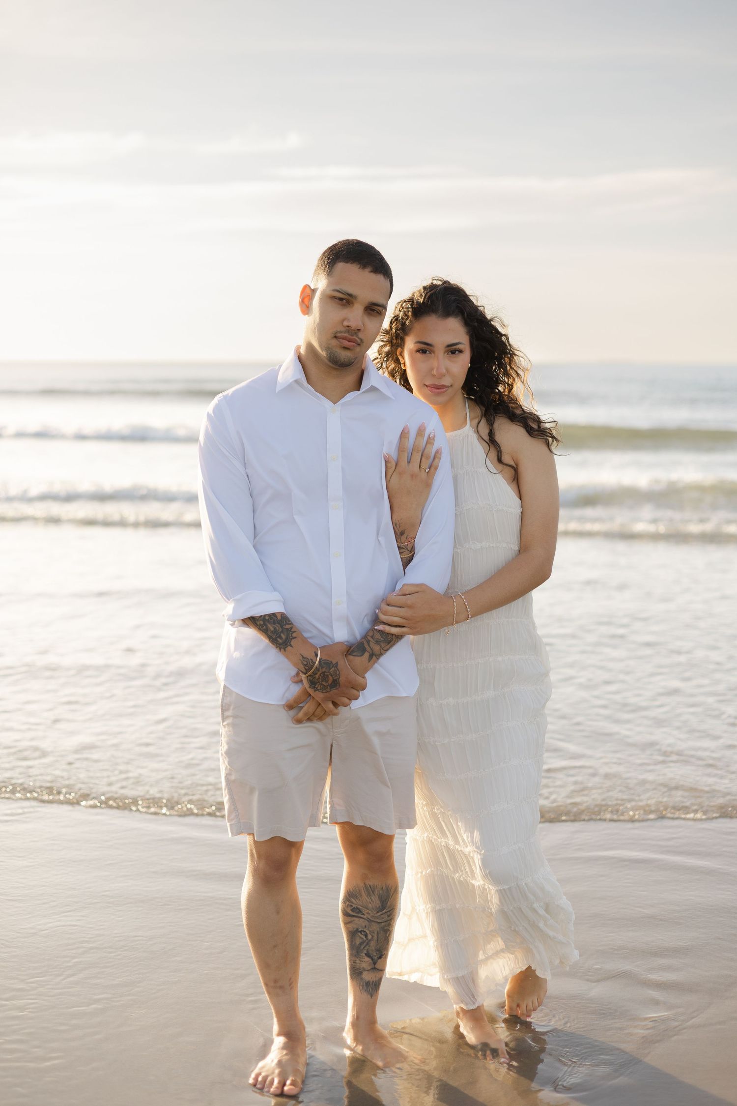 Engaged couple in white outfits standing together at ocean shore during golden hour.
