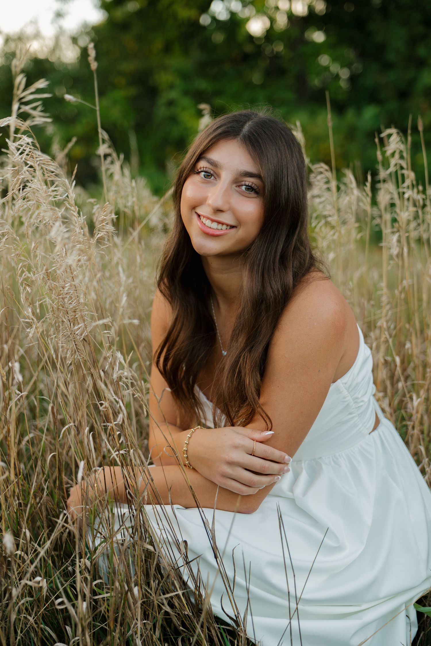 Young woman in white dress smiling among tall golden grasses with green trees in background.