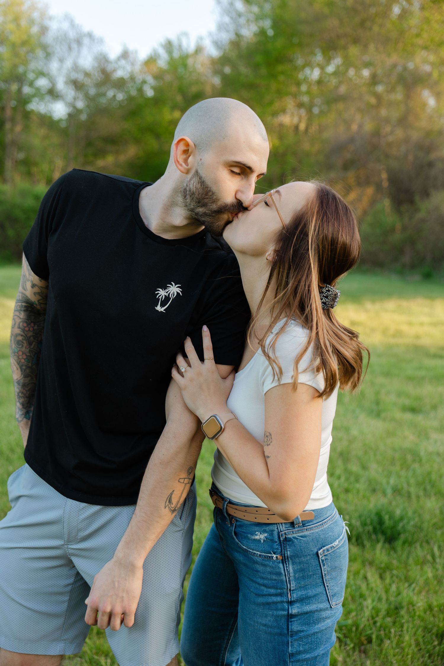Couple kissing outdoors in sunny green field, man in black tee and woman in white top sharing romantic moment.