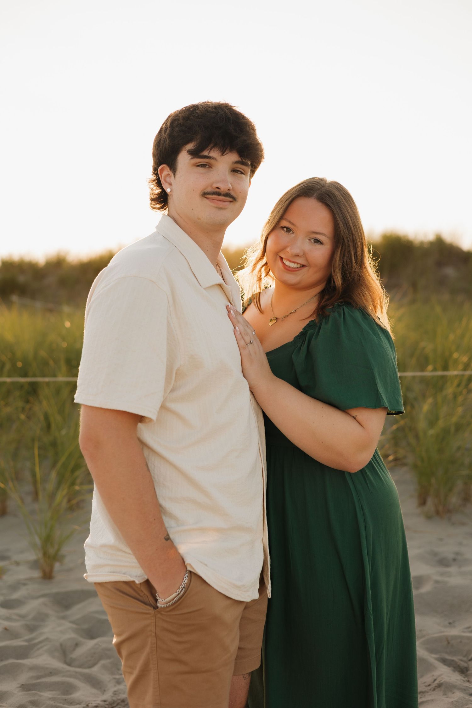 A young couple poses on a sandy beach at golden hour, woman in green dress leaning on man in white shirt.