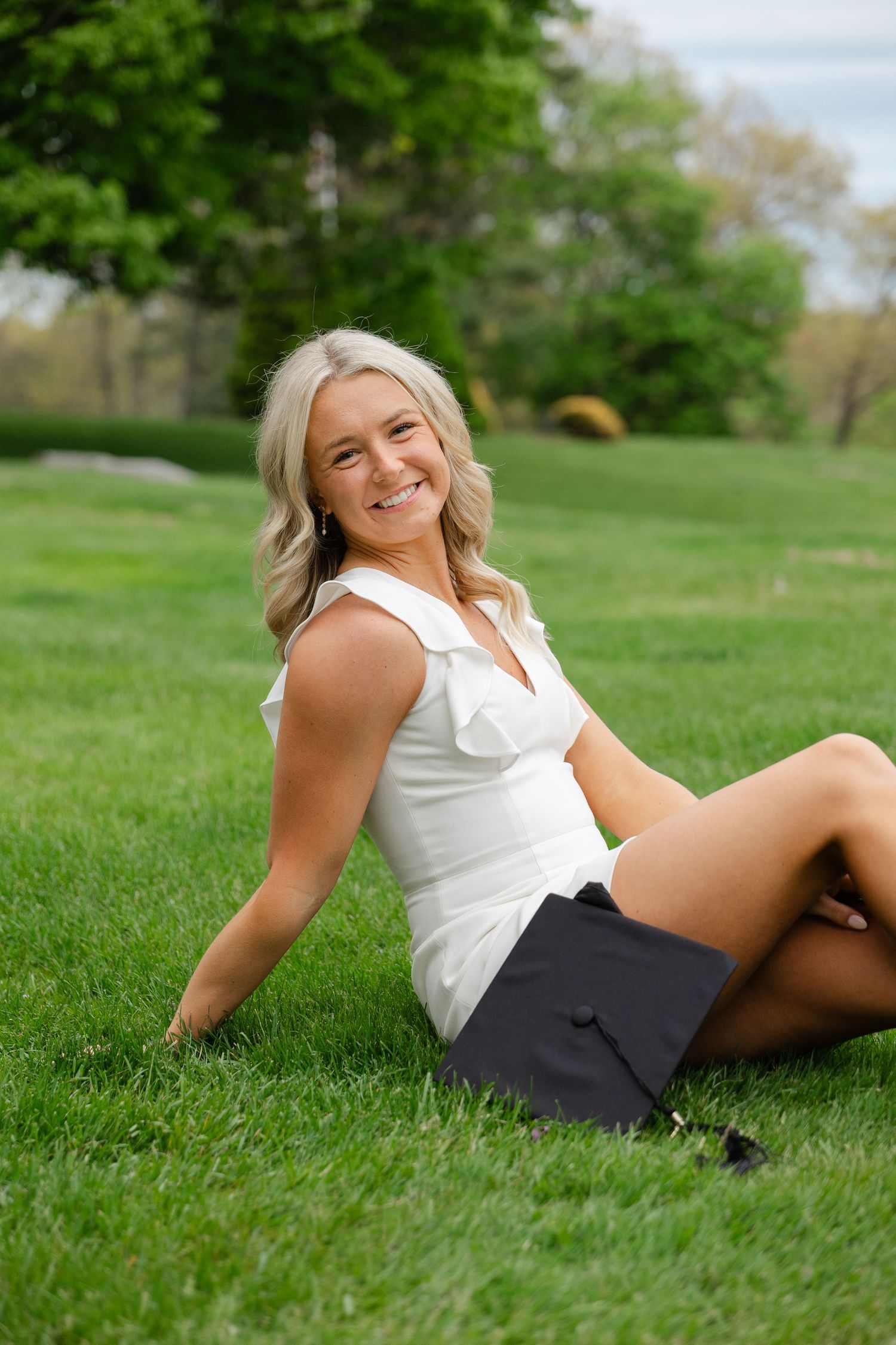 Smiling blonde woman in white top sitting on green grass holding a graduation cap outdoors.