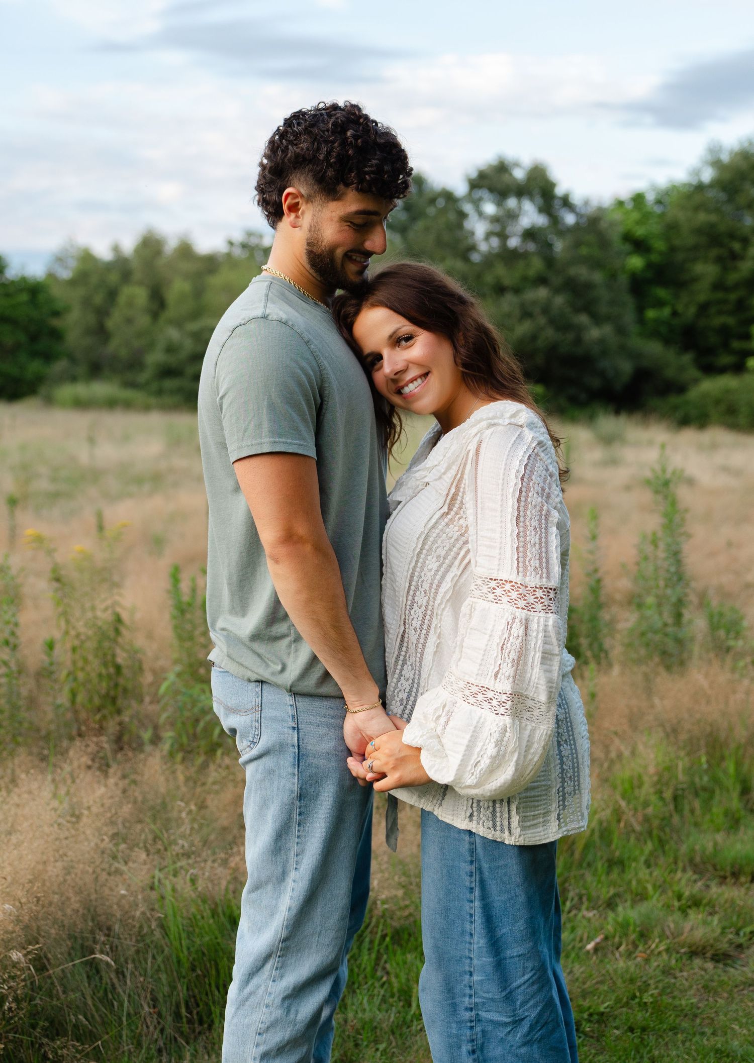 A couple holds hands and embraces in a lush green meadow, smiling warmly during an outdoor engagement photo session.