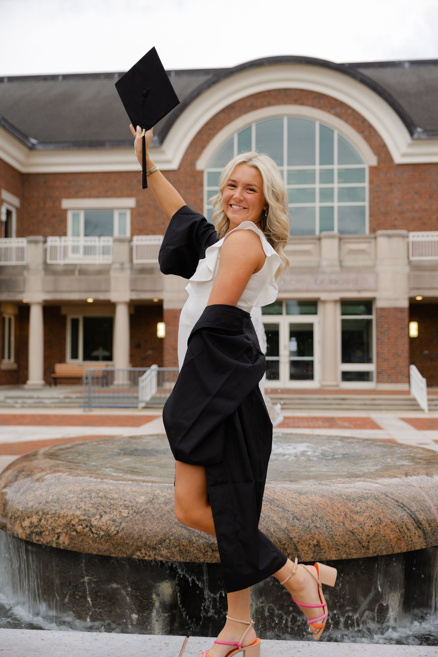 Joyful blonde graduate raising cap in air wearing black gown by brick university building.