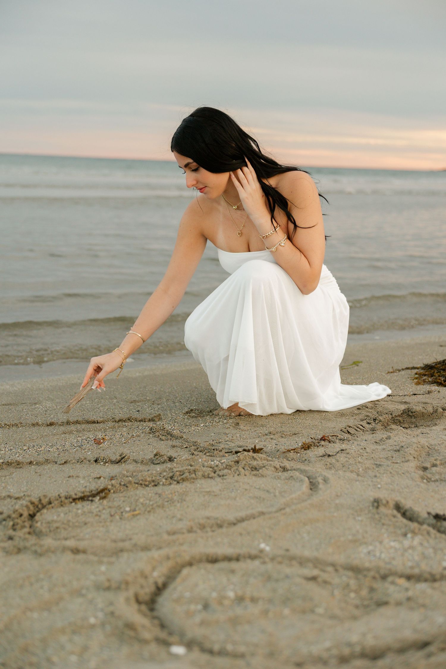 Woman in white dress writing in sand at beach during sunset.