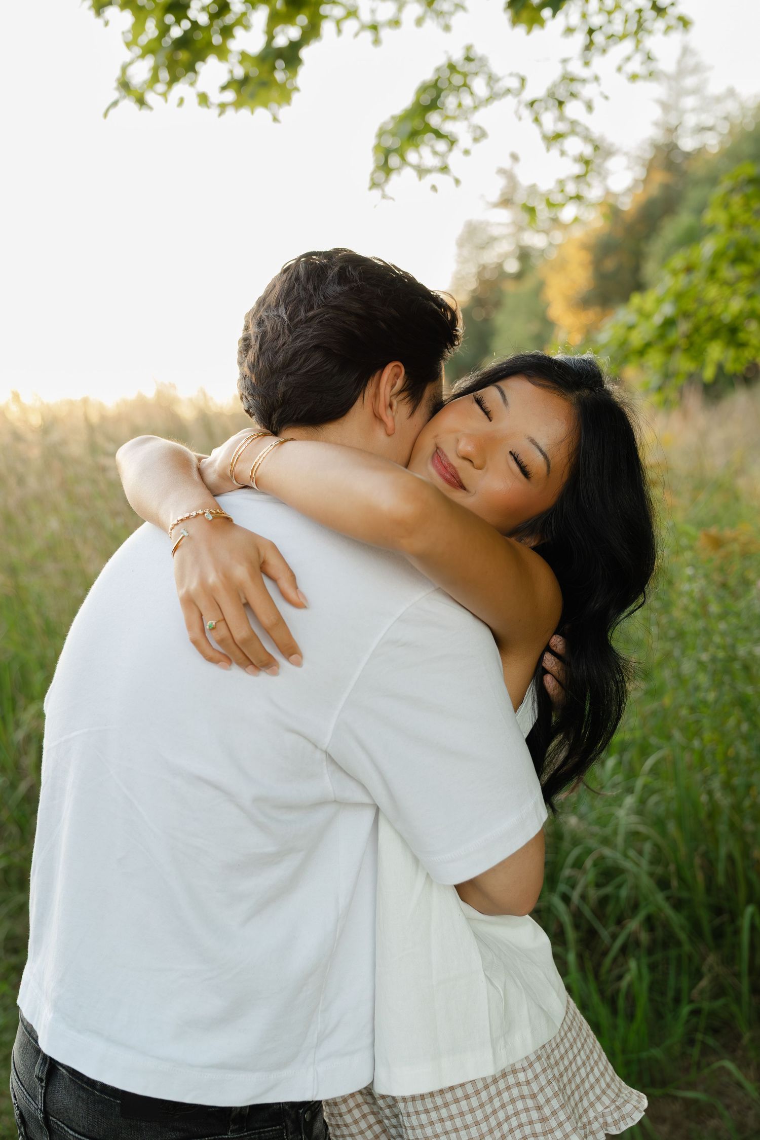 Couple embracing in a sunny green field, woman with dark hair hugging man in white t-shirt outdoors.