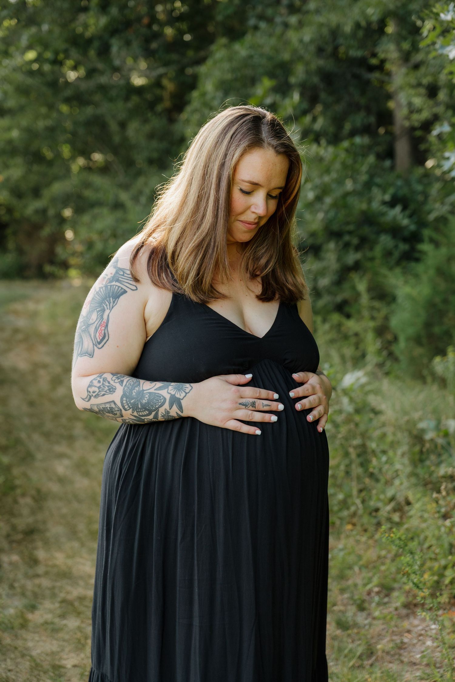 Pregnant woman in black dress holding baby bump outdoors, featuring tattoed arm, surrounded by lush green trees.