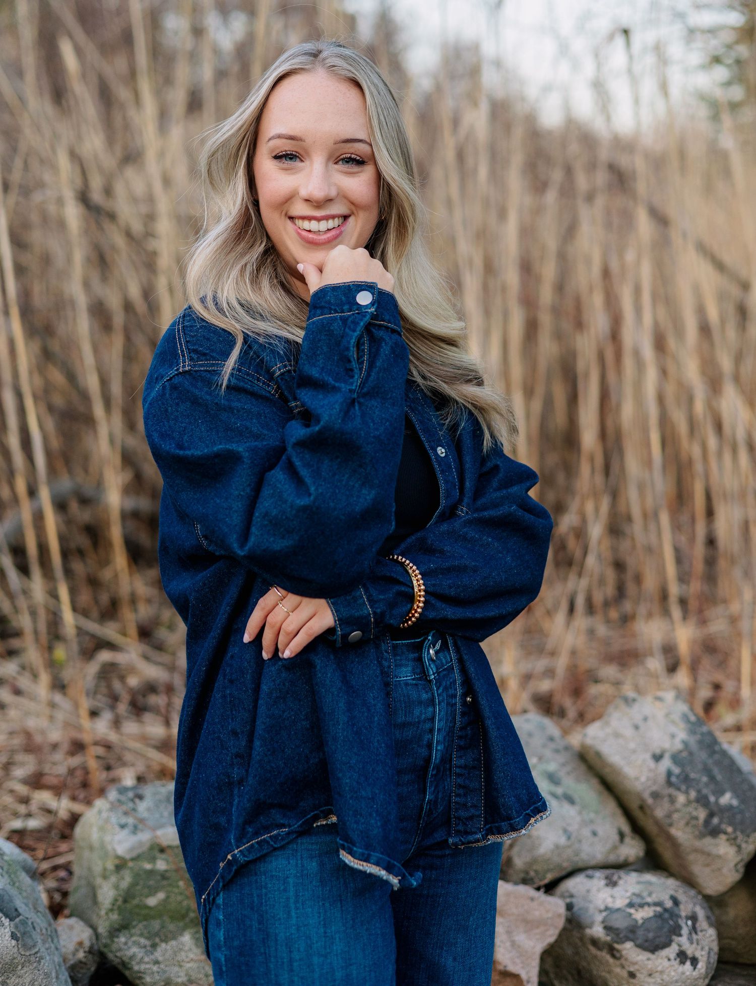Smiling blonde woman in oversized dark denim jacket posing outdoors near rocks and dried grass.