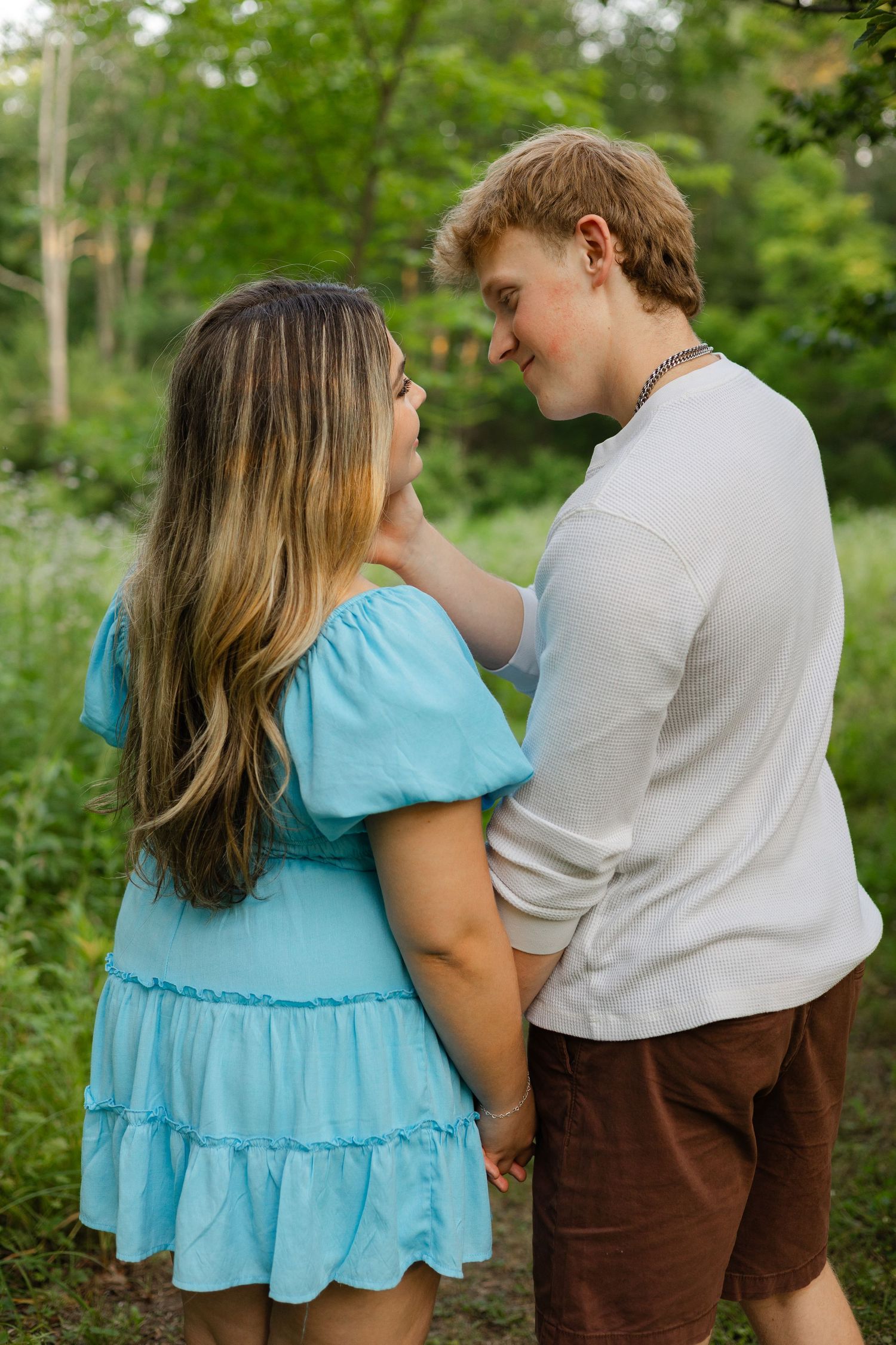 A young couple holds hands and gazes at each other in a lush green outdoor setting, she wears a blue dress.