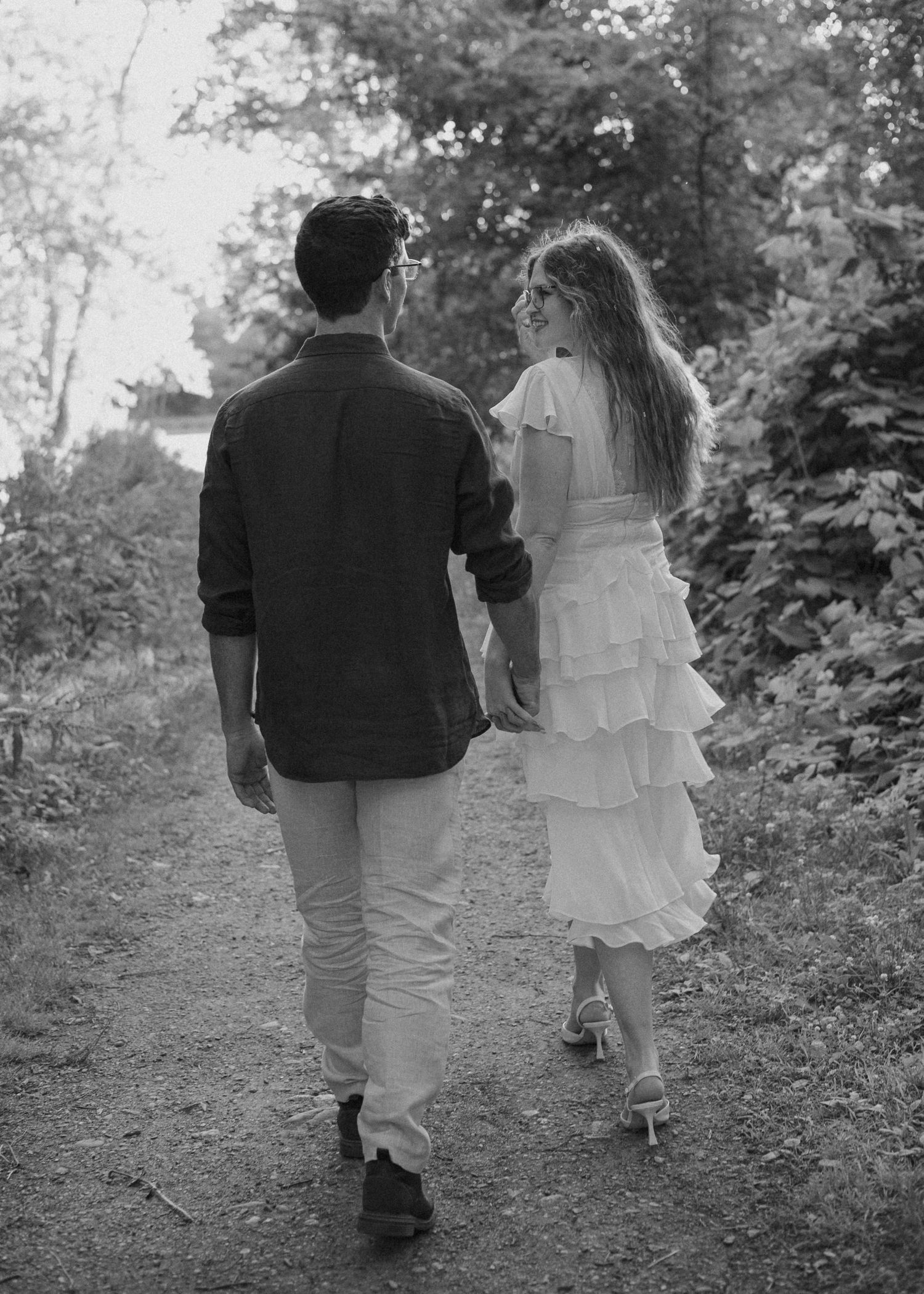 Black and white photo of a couple walking hand in hand down a forest path, woman in white dress.