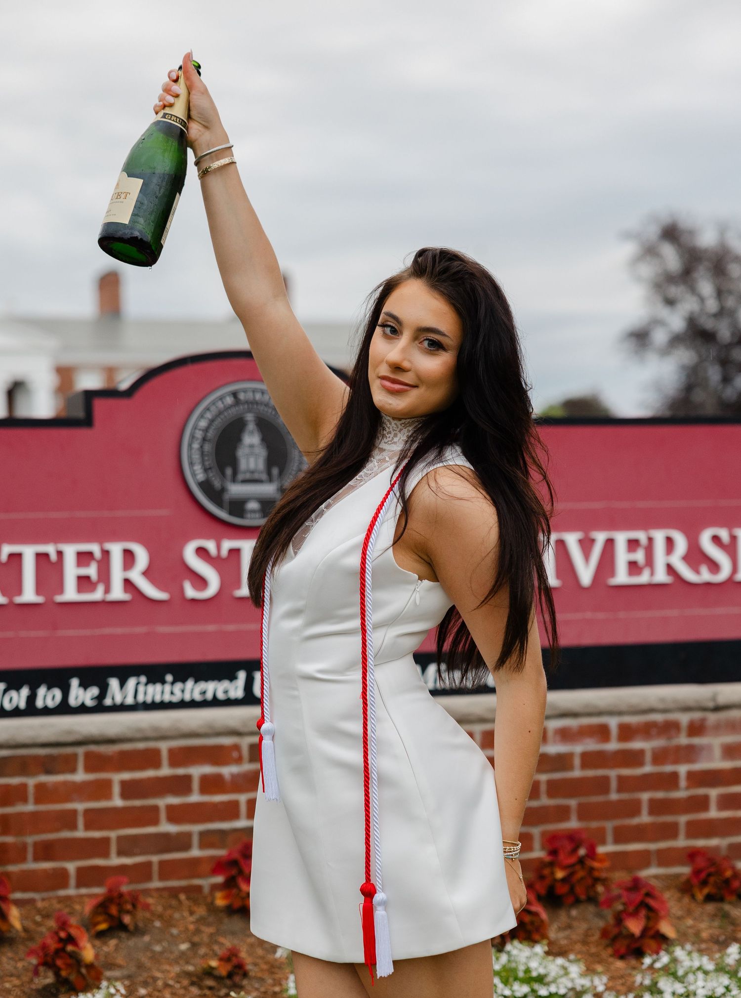 Graduate in white dress holds champagne bottle aloft in front of a university sign, wearing red graduation tassel.