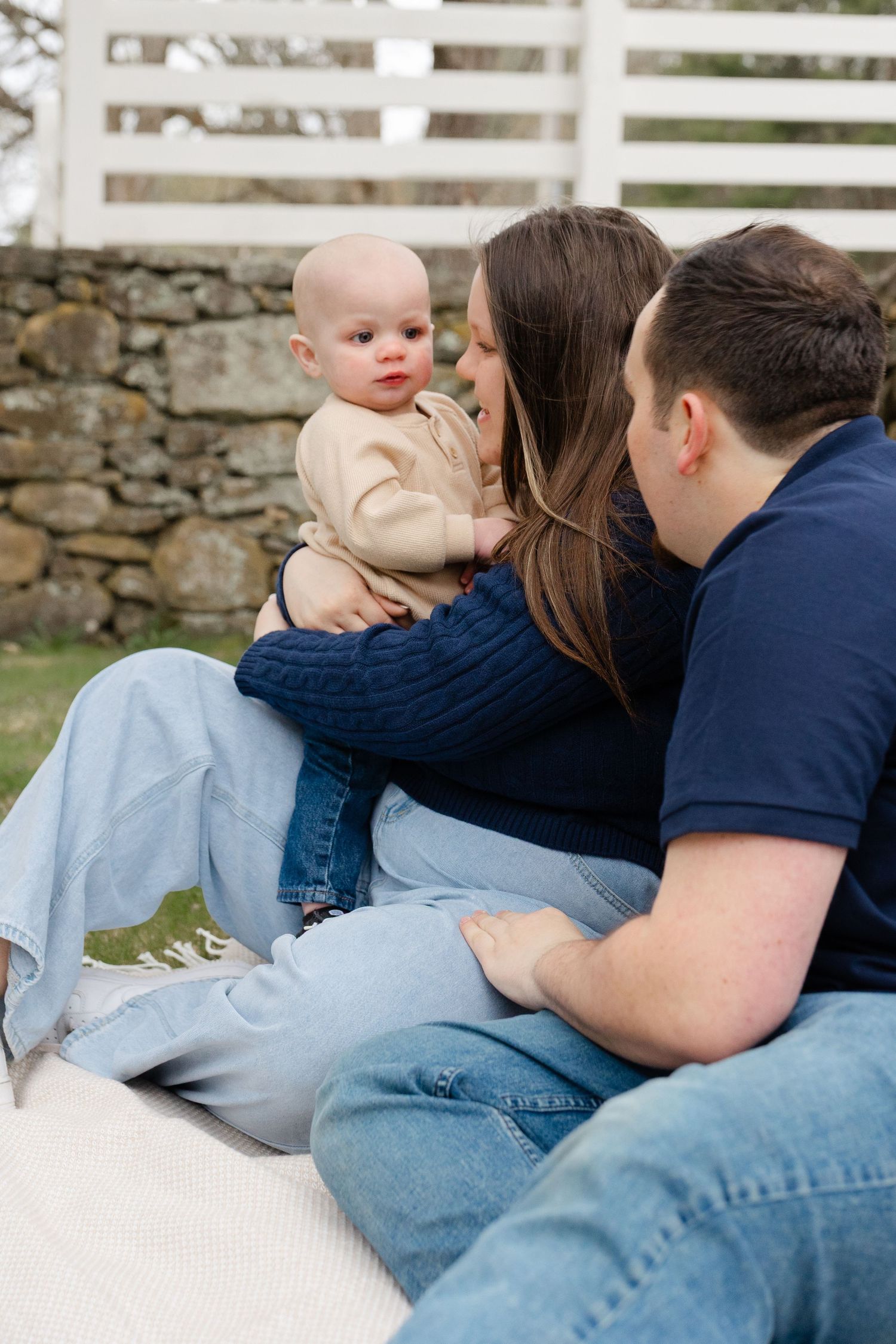 A young family sits outdoors on a blanket, parents lovingly holding their baby near a rustic stone wall.