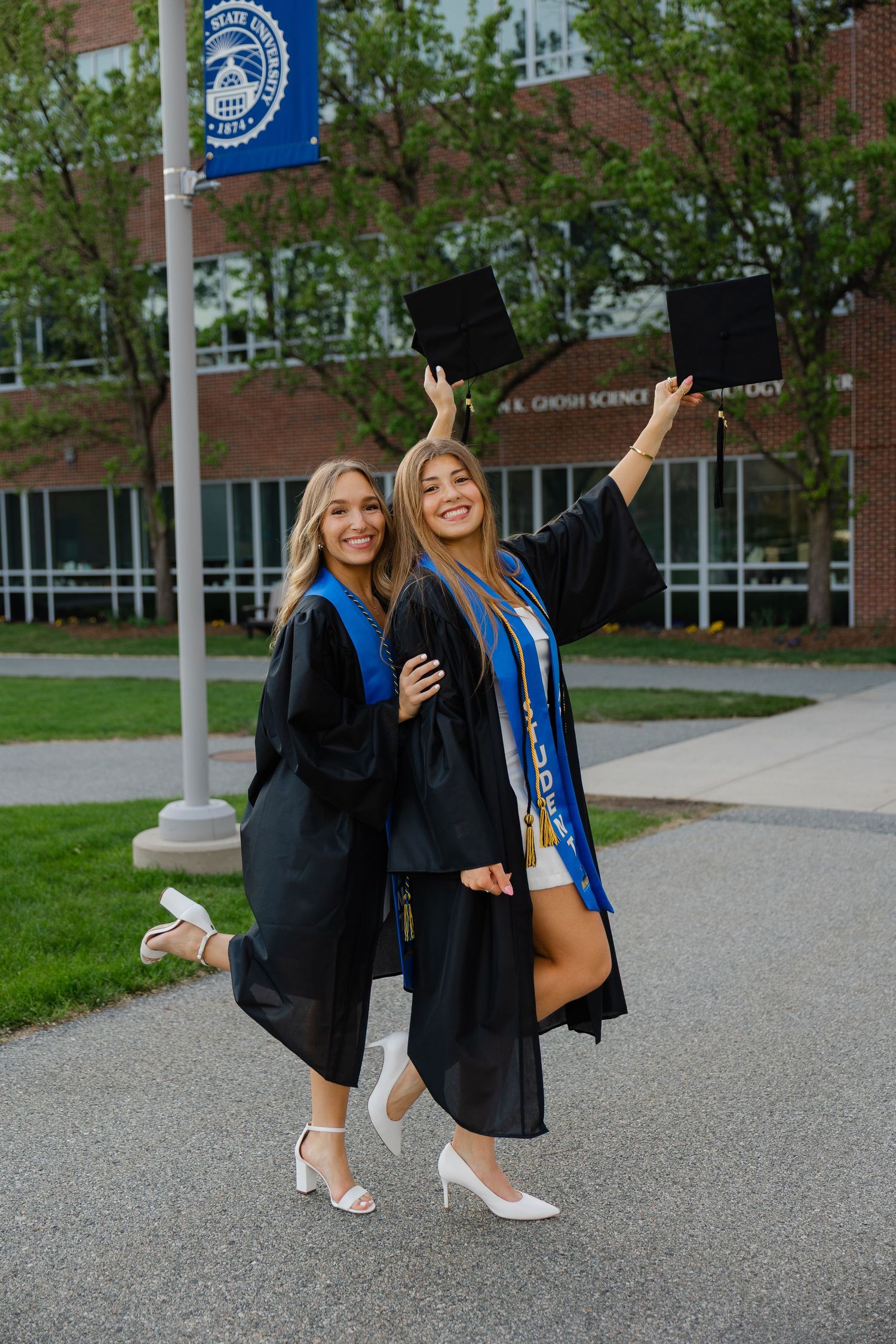 Two female graduates in black gowns and blue sashes celebrate outdoors, one tossing her cap in the air.