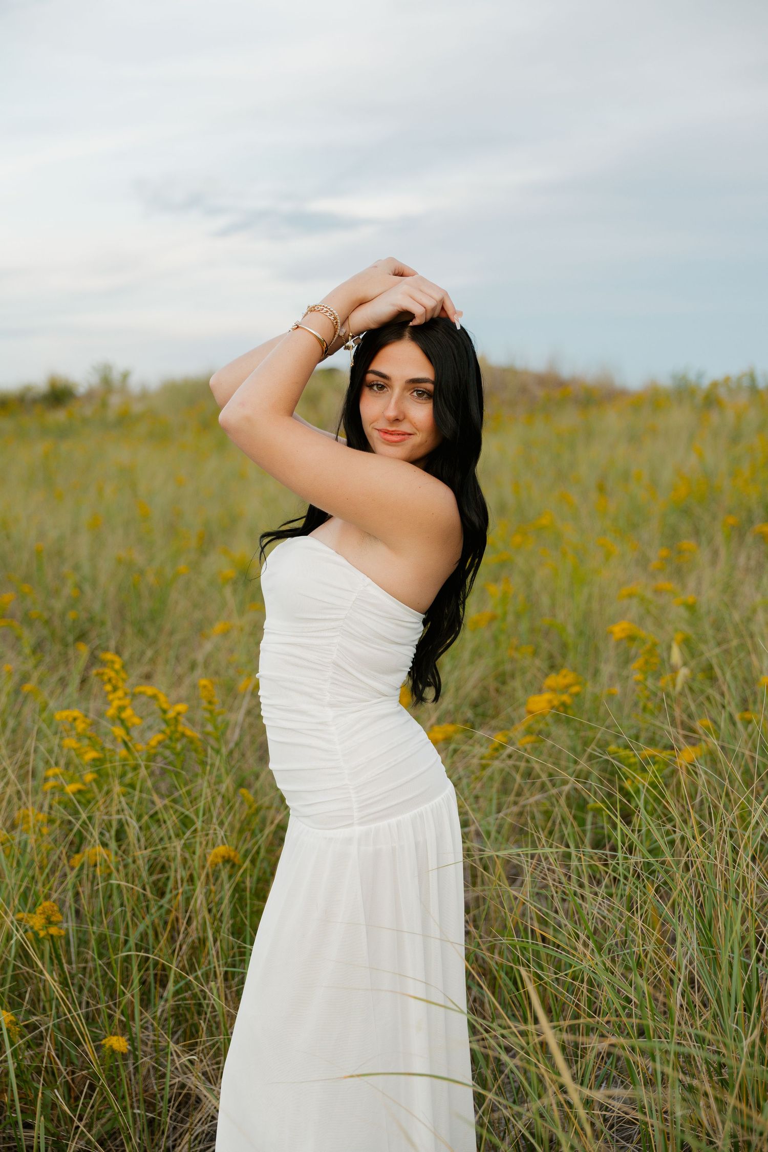 Young woman in white strapless dress posing in a wildflower field with arms raised, long dark hair, cloudy sky backdrop.