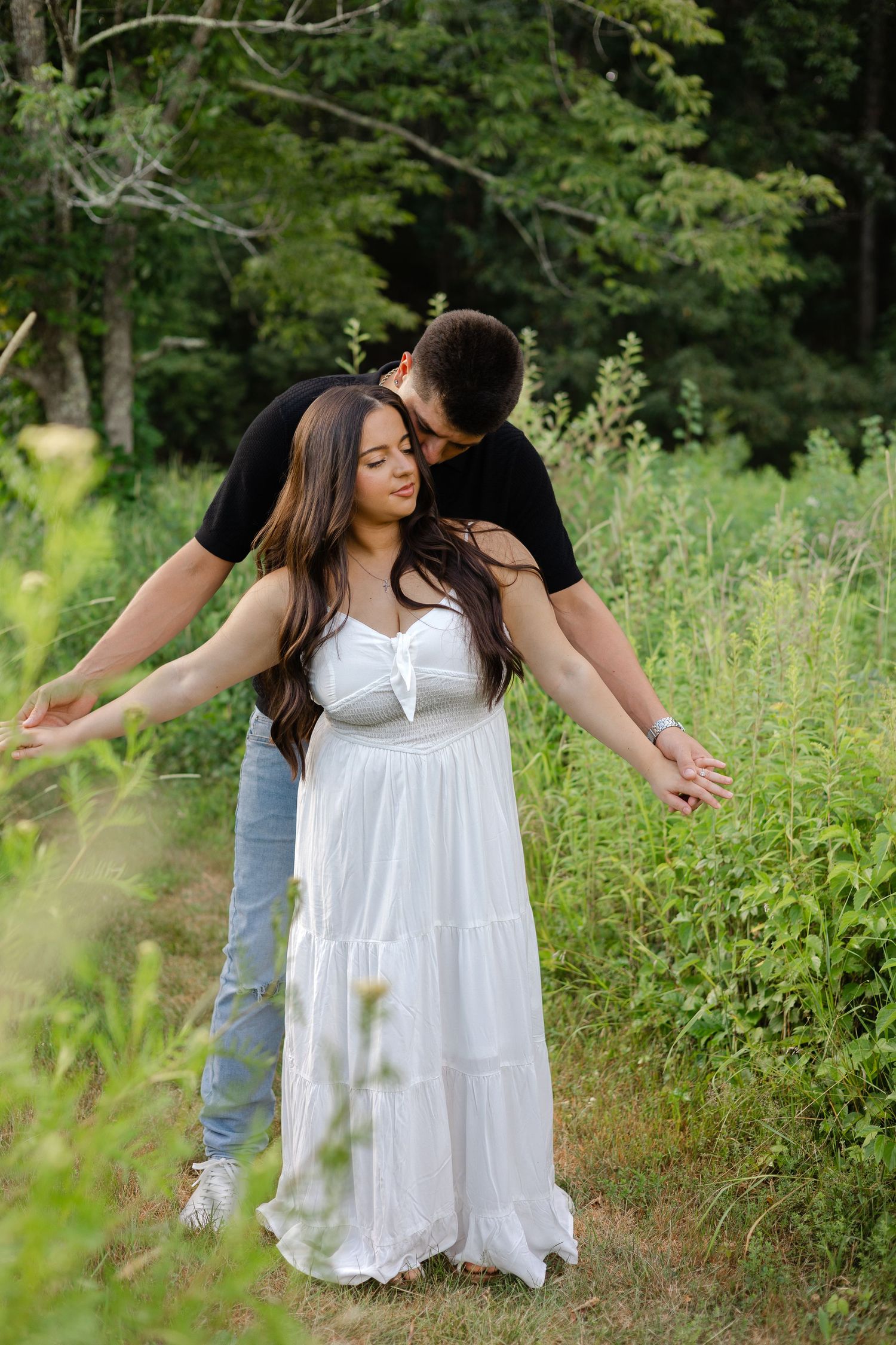 Couple in a lush green field, woman in white dress with arms out, man in black shirt kissing her head.