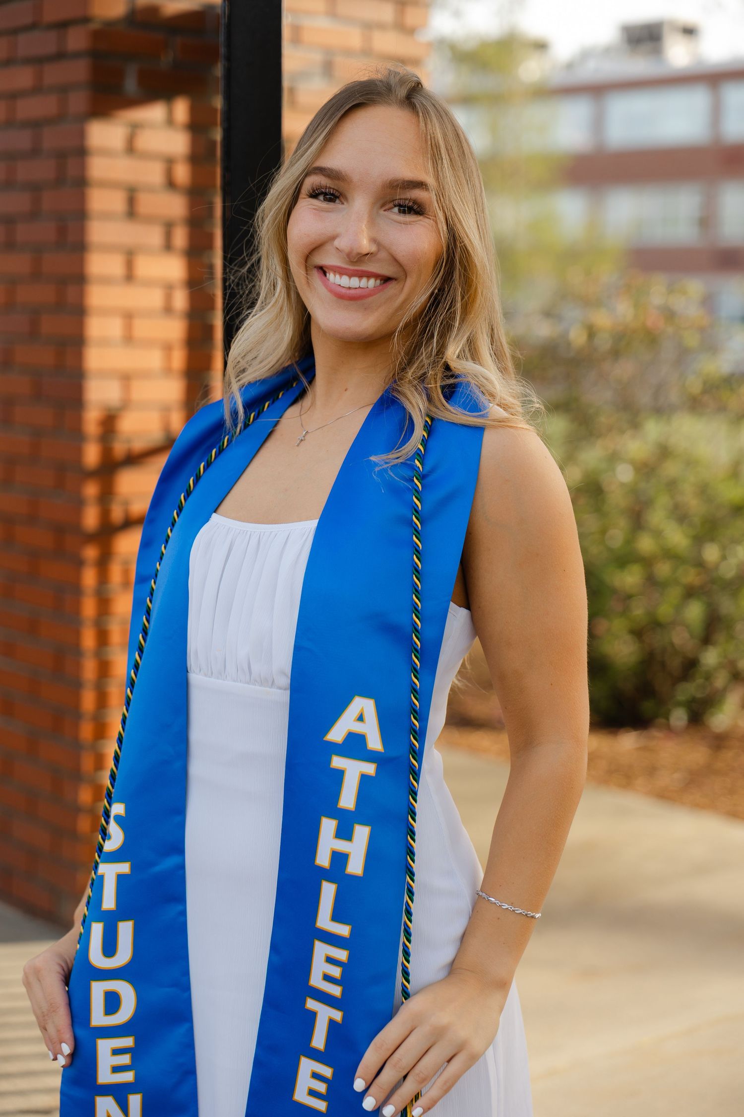 Smiling female graduate wearing a blue student-athlete sash over a white dress, posing outdoors near a brick building.