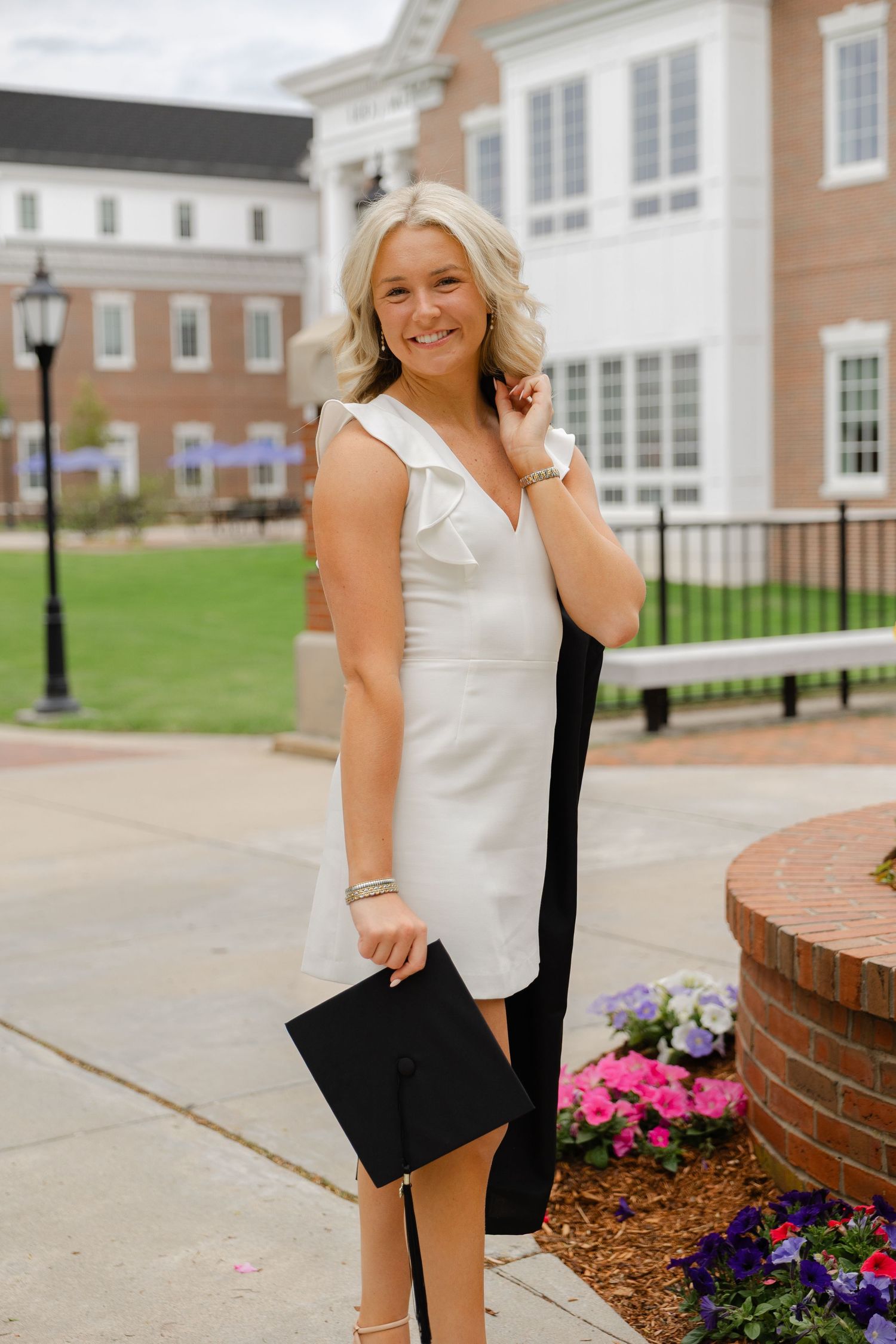 Smiling graduate in white dress holding cap near brick building with flowers.