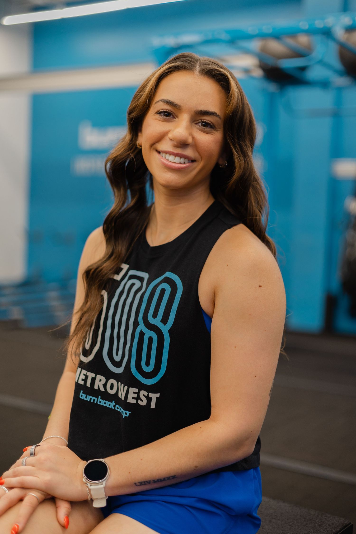 Smiling woman in a black 908 Metrowest tank top and blue shorts sits in a modern gym fitness studio.