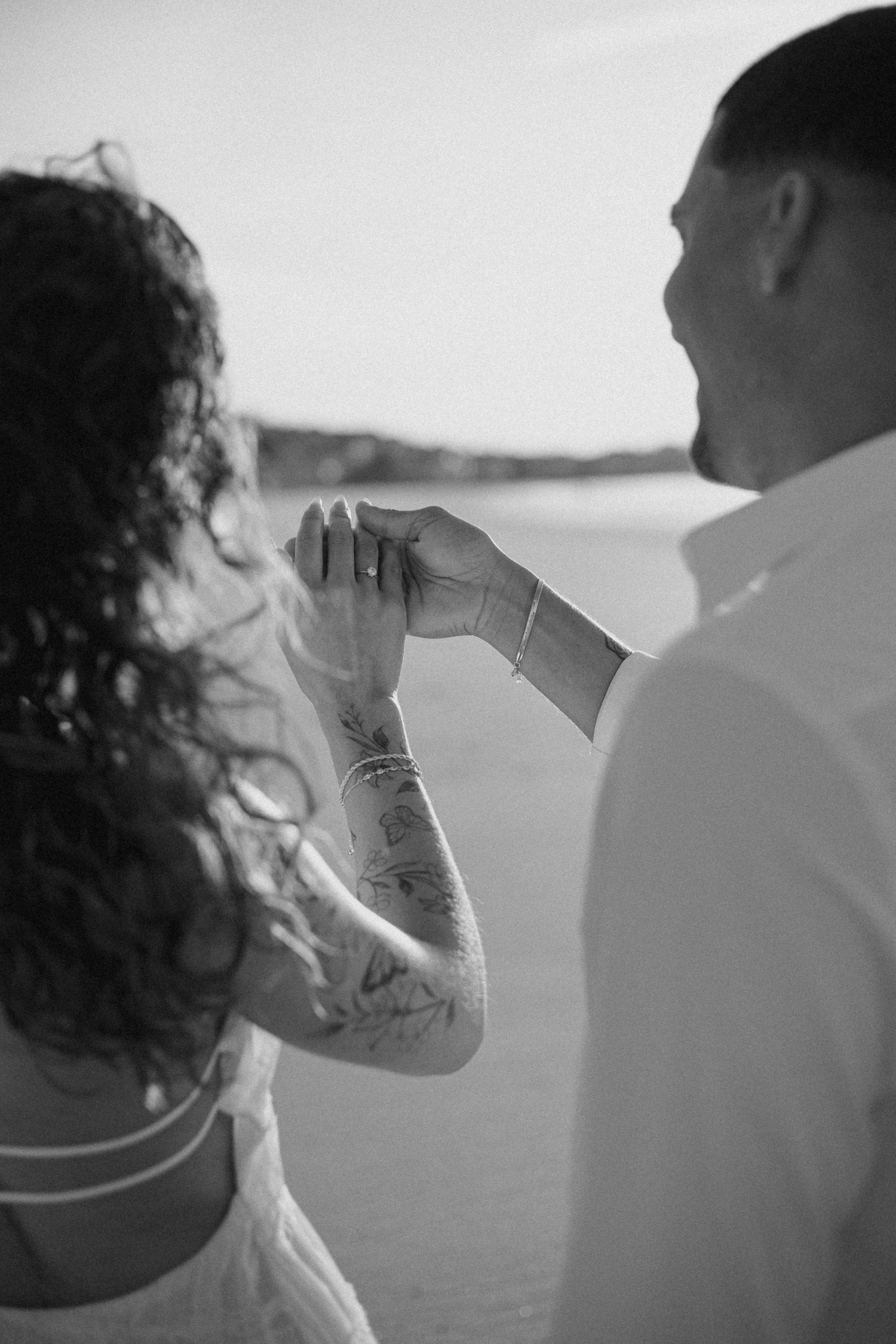 Black and white photo of a couple holding hands on a beach, woman with tattoos wearing a wedding ring.