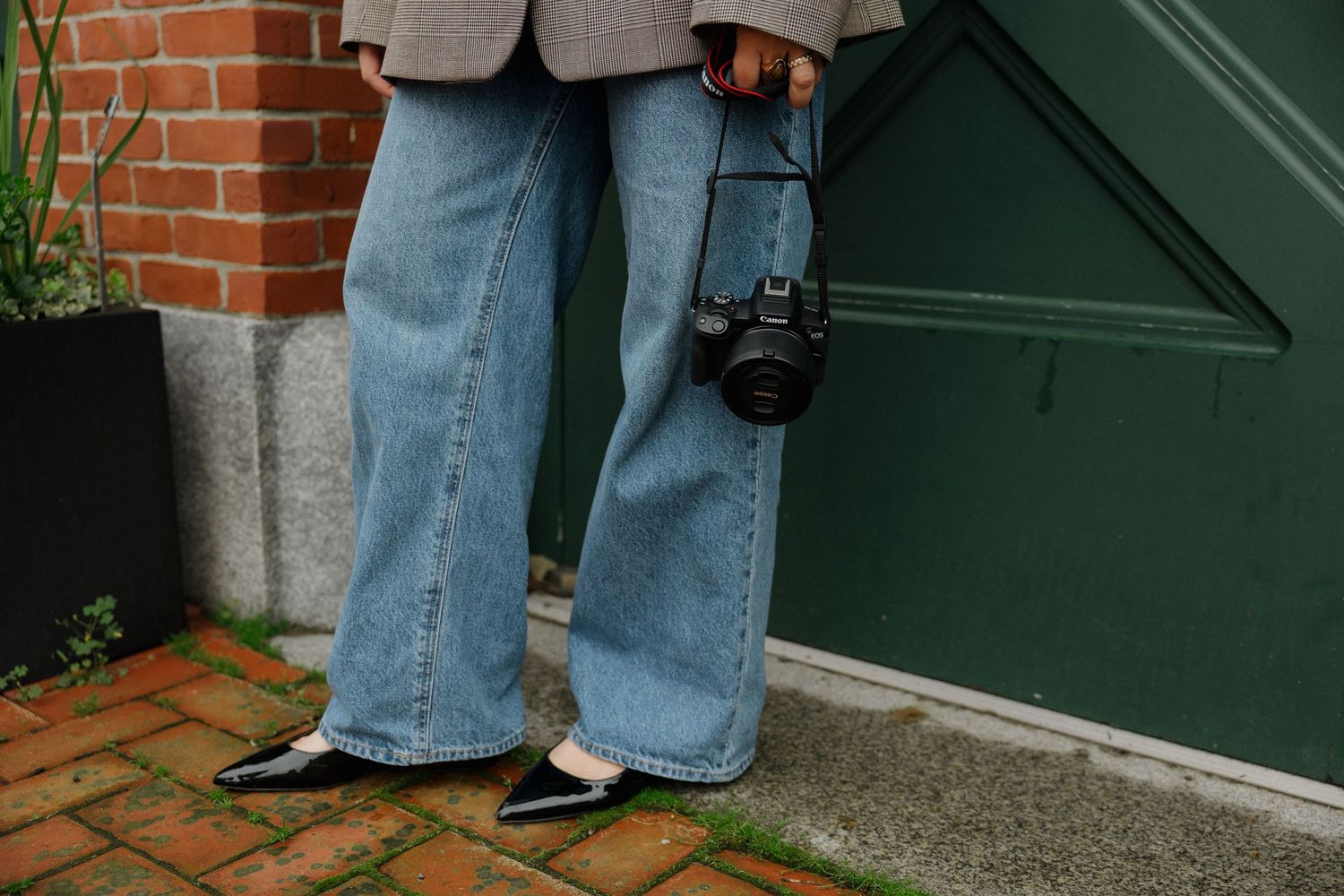 Person wearing wide-leg jeans and black flats holding a DSLR camera, standing near a green door and brick wall.